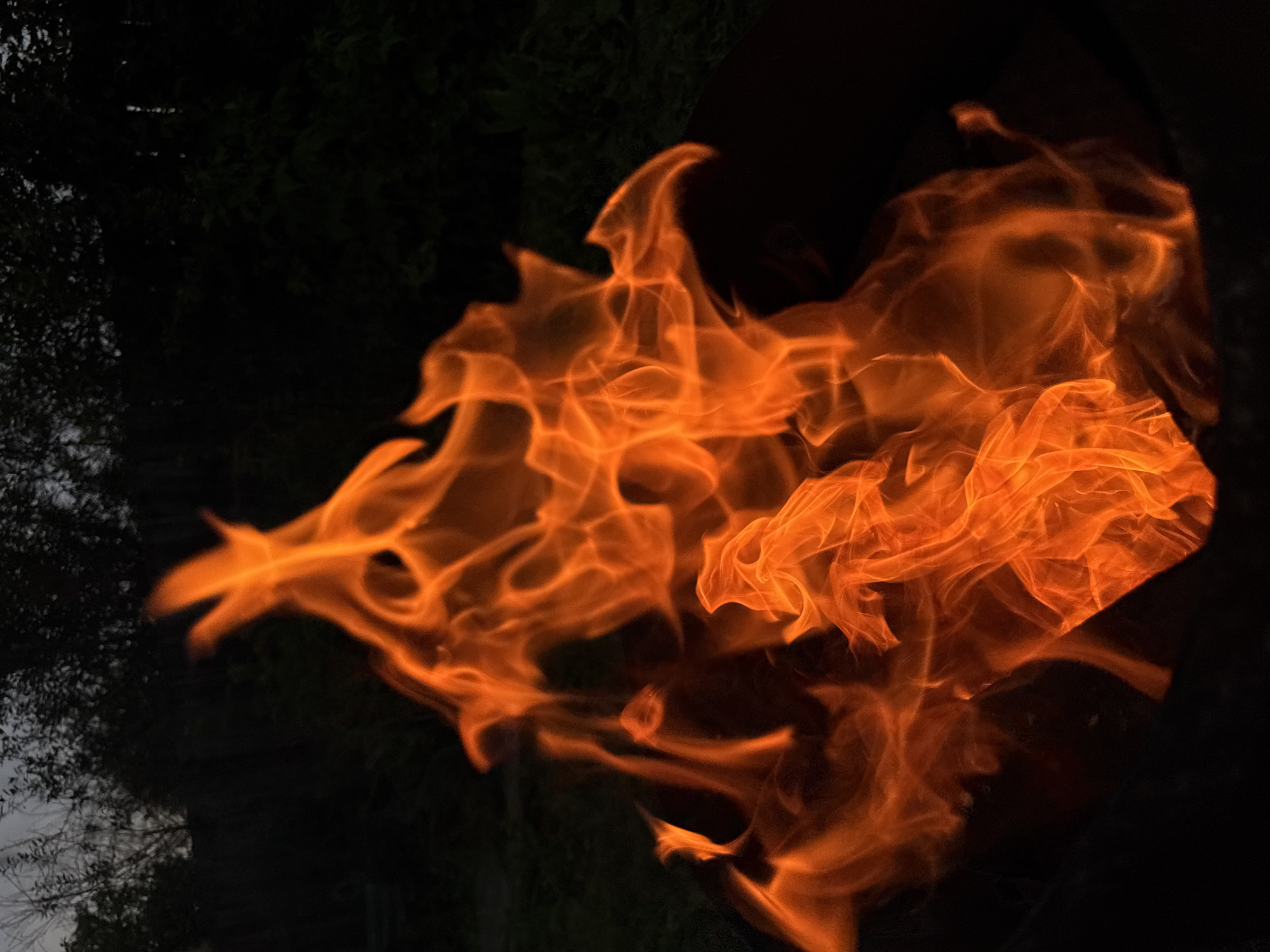 The image shows a close-up view of a fire burning in a dark setting. The flames are predominantly orange and yellow, with some areas appearing brighter and hotter than others. The background is dark, with hints of foliage or trees visible at the top, suggesting an outdoor location. The fire appears to be contained within a dark, circular object, possibly a fire pit or a barrel, which is partially visible at the bottom of the frame. There are no people or animals visible in the image. The overall impression is one of intense heat and light emanating from the fire against a dark, natural backdrop. The location in Chaikovsky, Russia is not visually discernible from the image itself; this information is provided as external context.