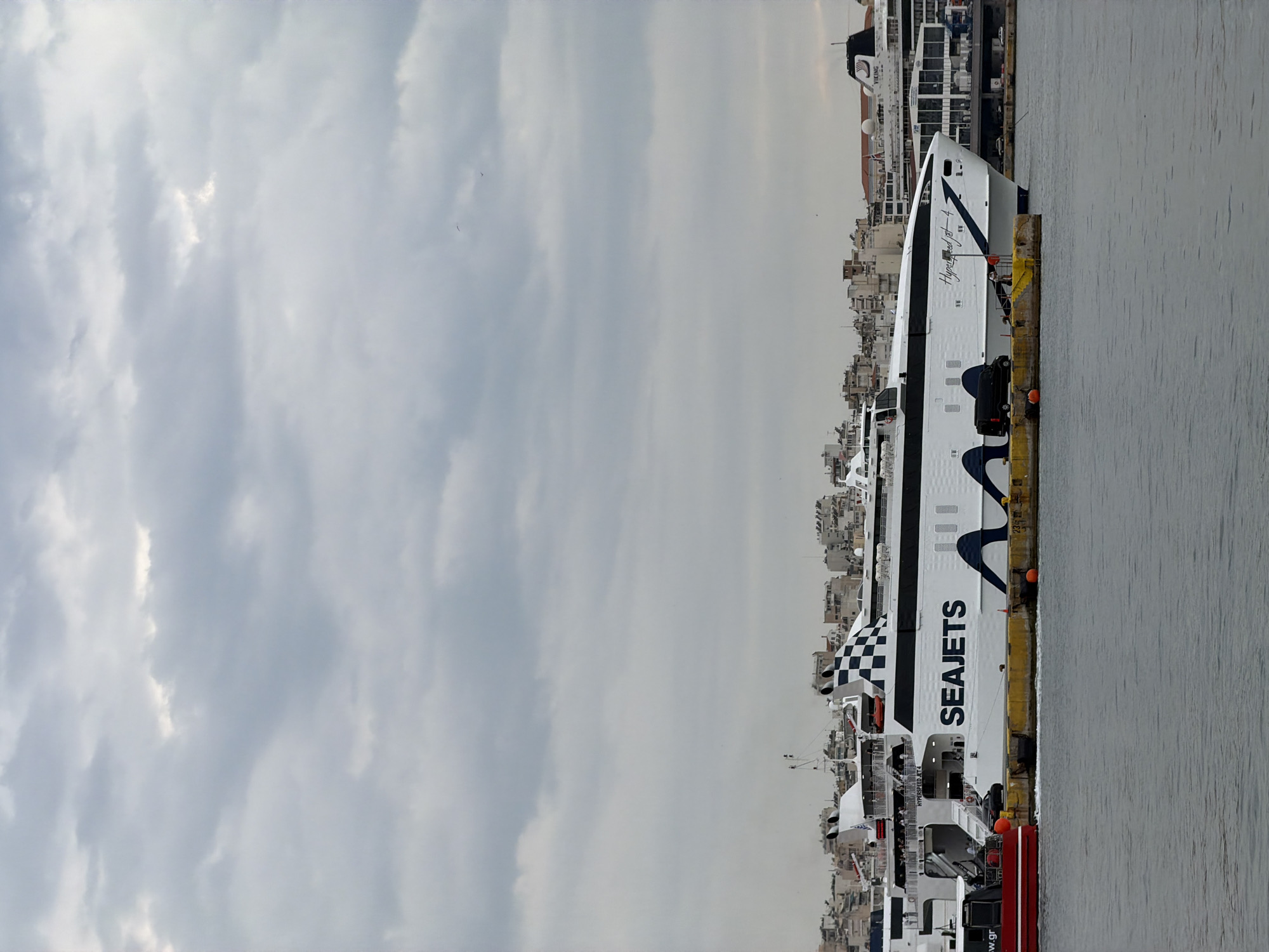 The image shows a large SEAJETS ferry docked at a pier in Piraeus, Greece. The vessel is white with dark blue and black accents, featuring prominent SEAJETS branding. Behind the ferry is the urban landscape of Piraeus, with buildings of varying heights. The sky is overcast. The water is calm. A person is visible on the boat.