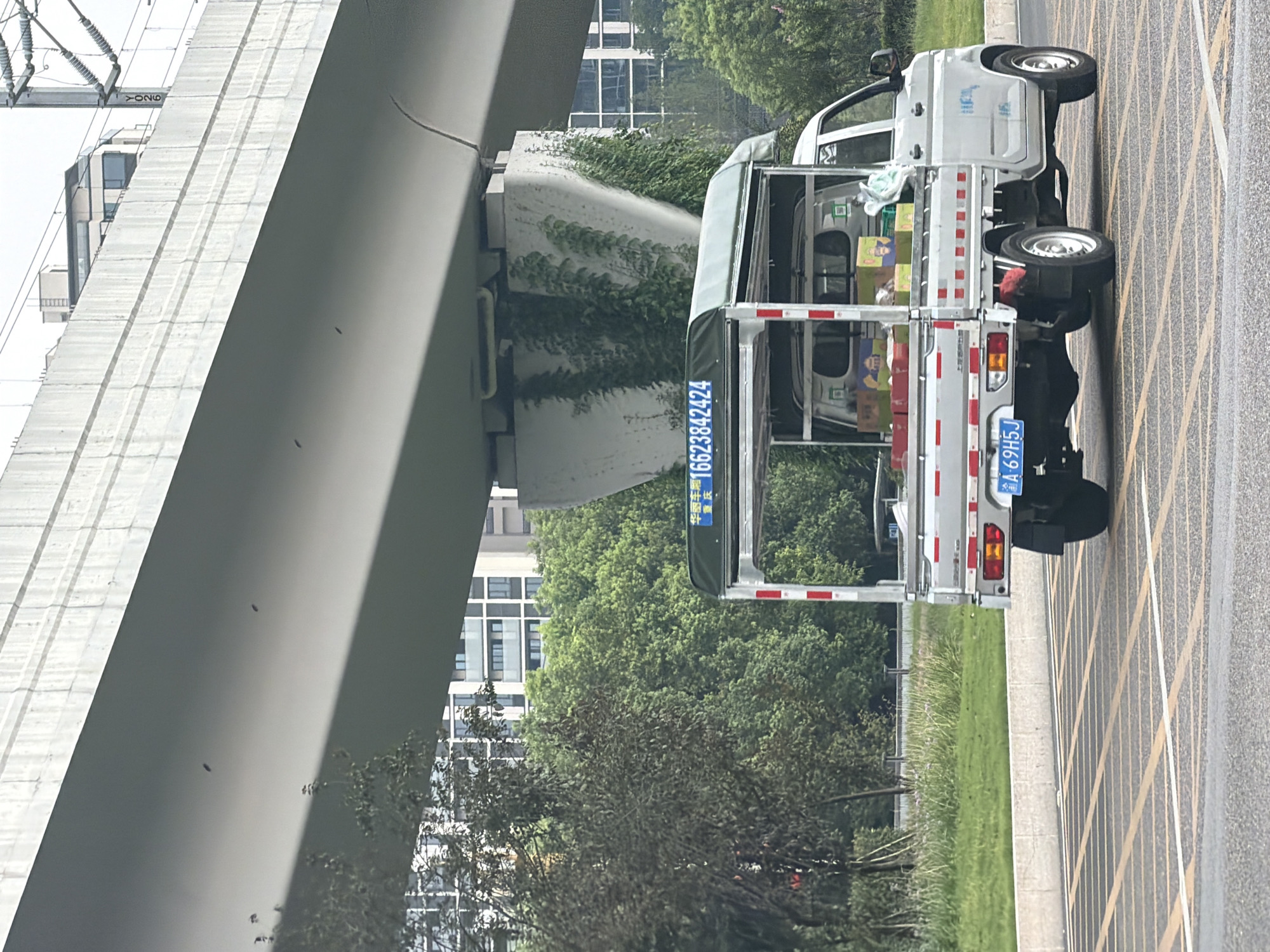 A silver, light-duty truck with an open cargo bed is parked on a road with yellow lane markings. The truck is loaded with boxes, some of which are green and yellow, and appears to be in motion or recently stopped. The truck has a license plate that reads "A·69H5J". Above the truck, a large concrete overpass casts a shadow. Behind the truck, lush green trees and modern buildings are visible, suggesting an urban environment. The scene appears to be in Jiangjin, China, as indicated by Chinese characters on a sign above the truck that read "华师车库" (Huashi Garage) and "16623842424".