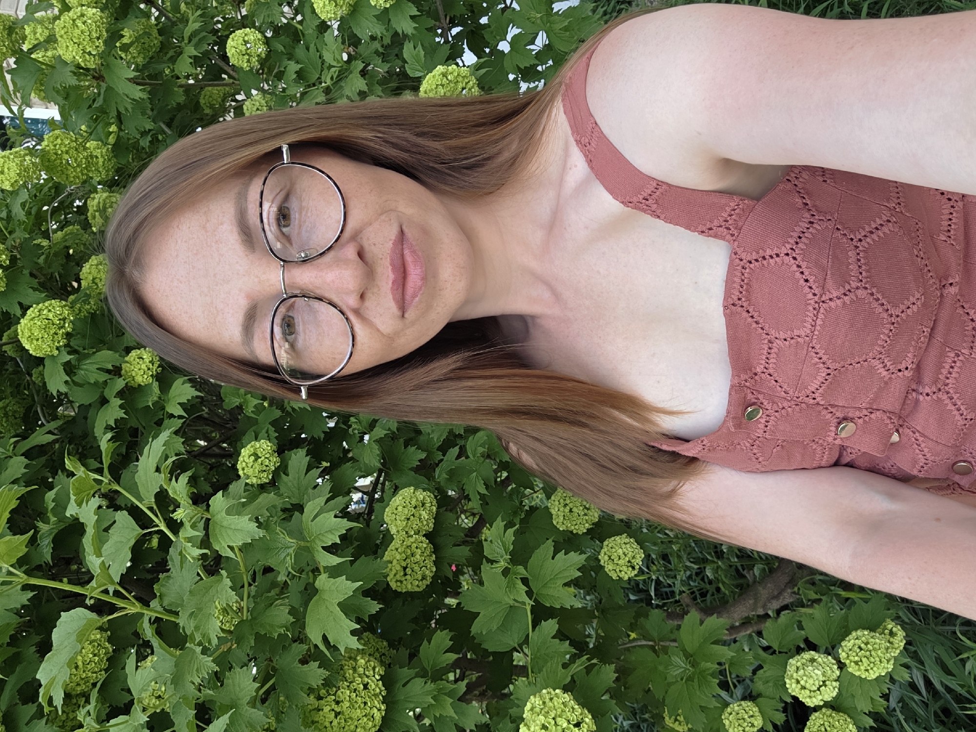 A woman with long auburn hair and wearing round glasses and a reddish-brown dress stands in front of a bush with green, spherical flowers. The background appears to be foliage, possibly a park or garden. The location is stated to be Chaikovsky, Russia.