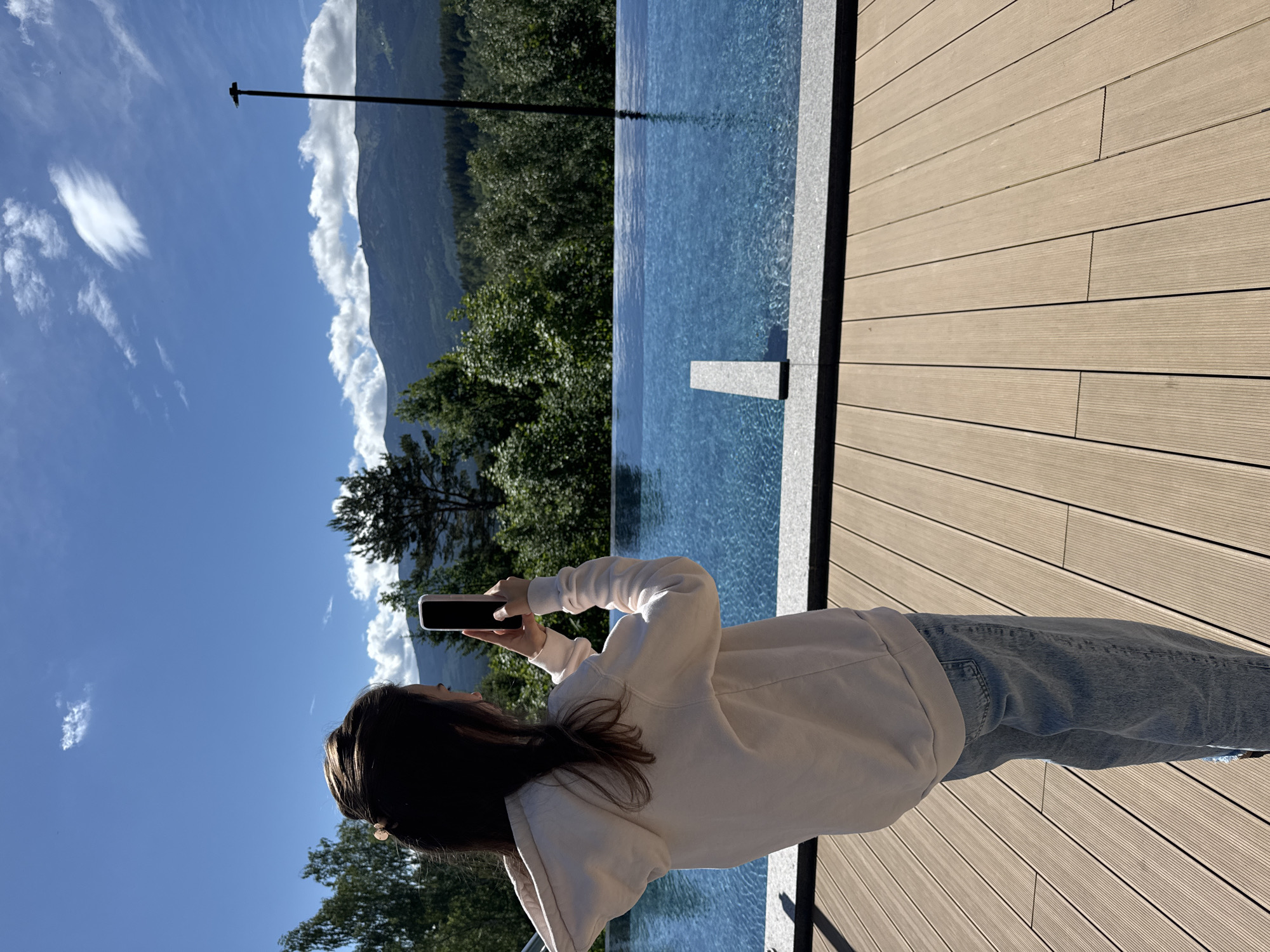 The image captures an outdoor scene featuring a person taking a photograph of a scenic landscape. The photo is oriented vertically, showing a person from the back, standing on a light-colored wooden deck next to an infinity pool. The person has long, dark hair and is wearing a light cream or white hooded sweatshirt and blue jeans. They are holding a black smartphone with both hands, pointing it towards the distant view, seemingly capturing the vista.
The infinity pool, filled with clear blue water, stretches across the midground, reflecting the sky and surrounding elements. Beyond the pool, a dense forest covers rolling hills, leading up to a range of mountains in the background. The sky is a vibrant blue with scattered white, wispy clouds. A tall, slender pole with a small horizontal element at its top stands upright near the edge of the pool, silhouetted against the bright sky and mountains.
The setting appears to be a luxurious vantage point, possibly at a resort or private residence, offering expansive views of nature. The bright daylight suggests a sunny day, likely mid-morning or afternoon, with clear weather. There is no visible text in the image.