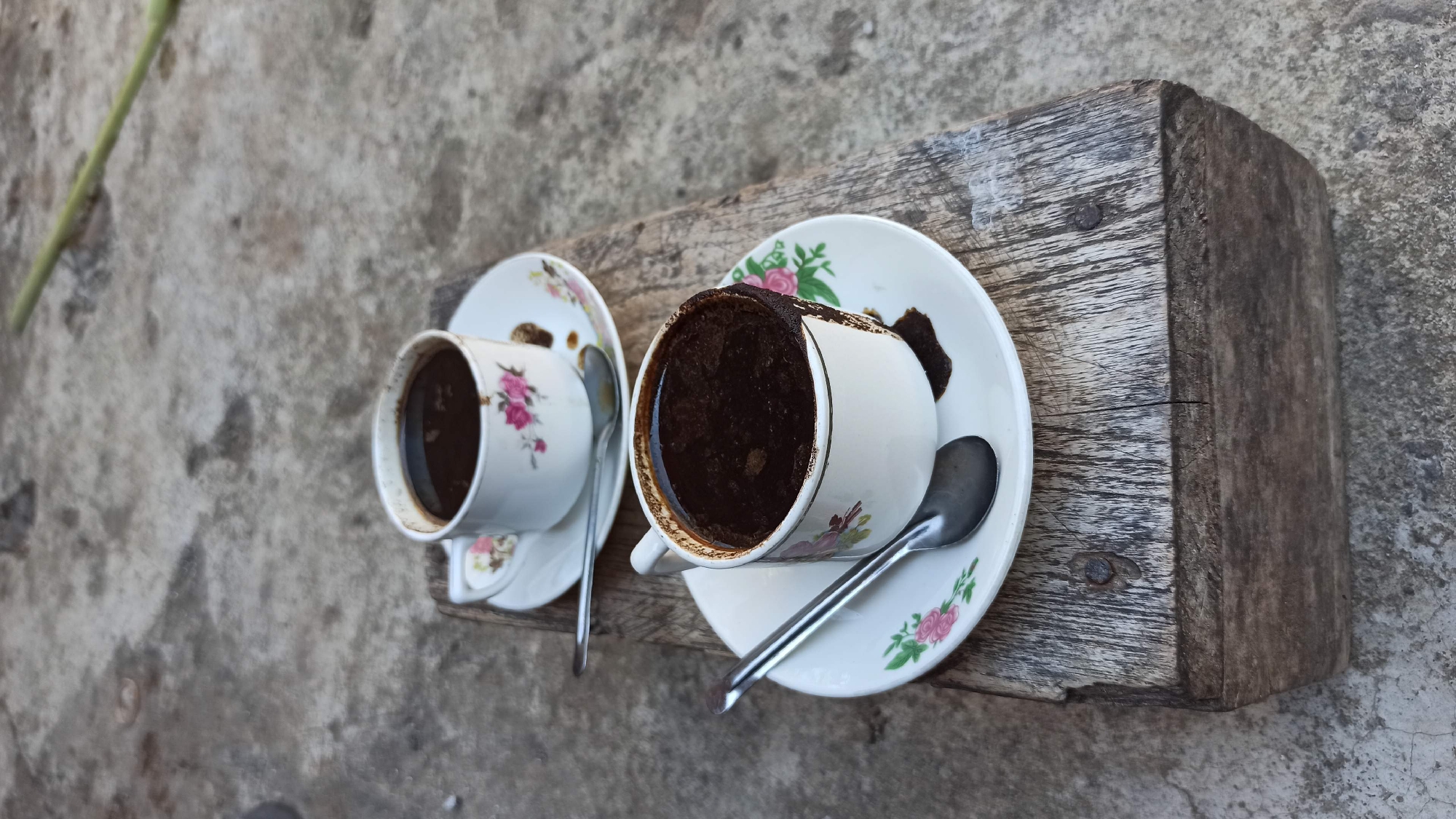 Two cups of black coffee, each with a small spoon and saucer, are placed on a rustic wooden surface. The cups are white with a floral design featuring pink and green flowers. One cup is slightly fuller than the other, with some coffee spilled down the side. The wooden surface appears weathered and is placed on a concrete or stone ground. A thin green stick is visible in the blurred background. The image was taken in Gegeran, Indonesia.
