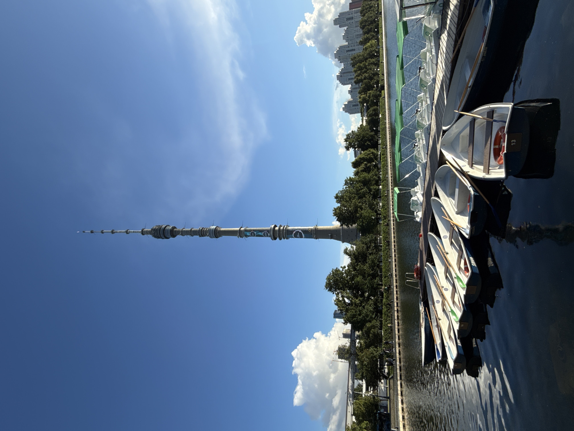 The media file depicts a view of the Ostankino Tower in Moscow, Russia, from a waterfront. The tower, a prominent landmark, stands tall against a clear blue sky with scattered white clouds. In the foreground, several small boats are docked along a wooden pier. The boats are mostly white with some dark hulls and appear to be rowing boats, some equipped with oars. One of the boats has a life preserver with the number "13" visible.
The scene is bathed in bright sunlight, suggesting it is daytime, likely late morning or afternoon. Lush green trees line the edge of the water, creating a natural barrier between the waterfront and a distant cityscape of buildings. The water is dark and reflective, mirroring the sky and the boats. The overall atmosphere seems peaceful and serene, ideal for recreational activities on the water.
The composition of the image is vertical, with the tower dominating the left side and the boats and pier occupying the right. The perspective is from slightly below the tower, emphasizing its height. No people are clearly visible, suggesting the area is either not very busy or the focus is on the scenery and the docked boats. The weather appears to be fair and sunny, with a gentle breeze implied by the calm water surface.