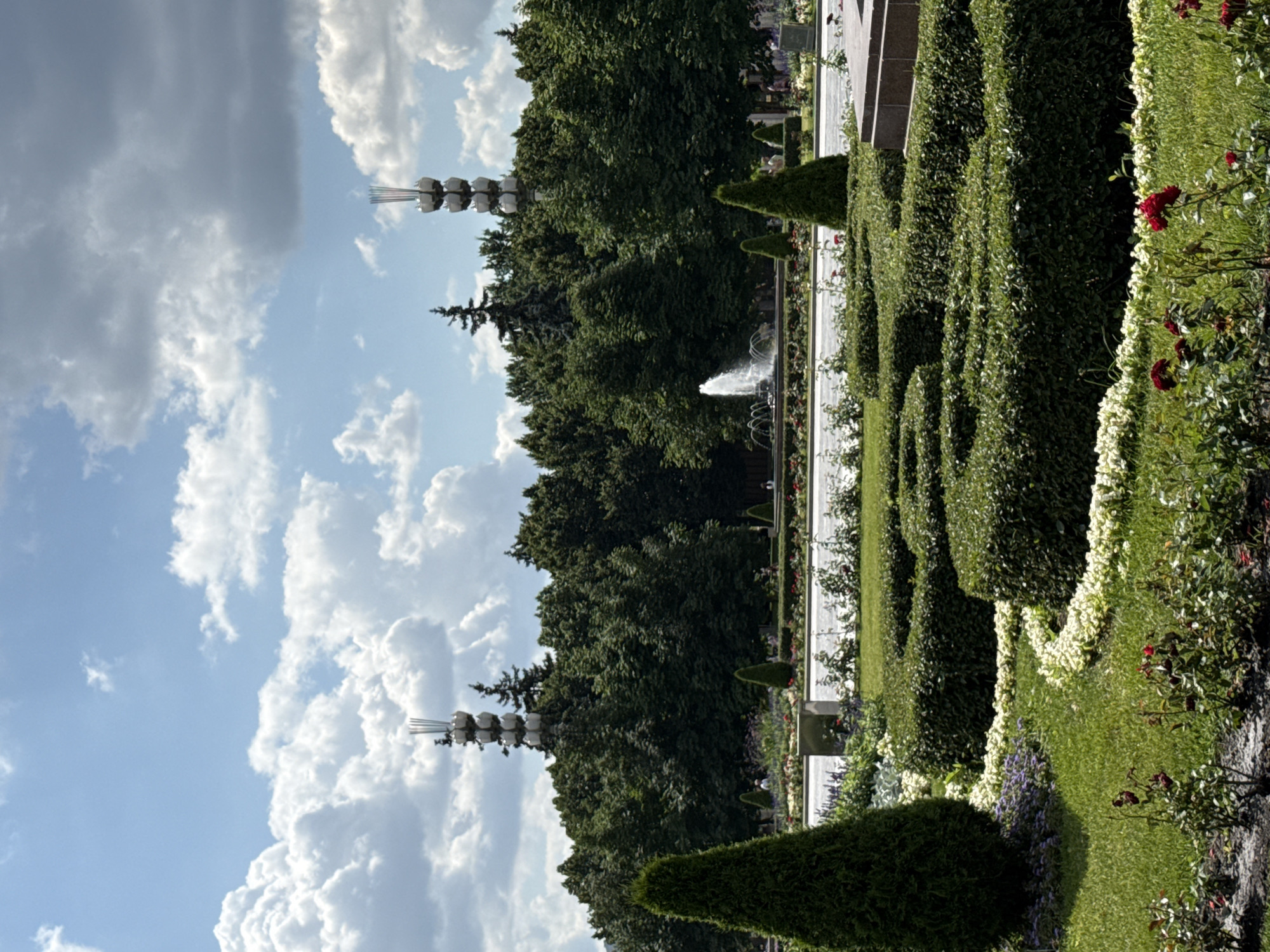 The image depicts a summer day in Ostankino Park in Moscow, Russia. In the foreground, a meticulously landscaped garden features manicured hedges arranged in a maze-like pattern, bordered by white flowers and green grass. Several vibrant red roses are in bloom. A tall, conical evergreen tree stands to the left.
In the mid-ground, a wide fountain sprays water into the air, and further back, a line of people can be seen near a walkway and more garden beds. Beyond the trees, two identical, distinctive radio towers with a stacked, spherical design and antennae at the top are visible against a backdrop of lush green trees. The sky is bright blue with scattered white, puffy clouds. The overall atmosphere is one of a well-maintained urban park on a sunny day.
