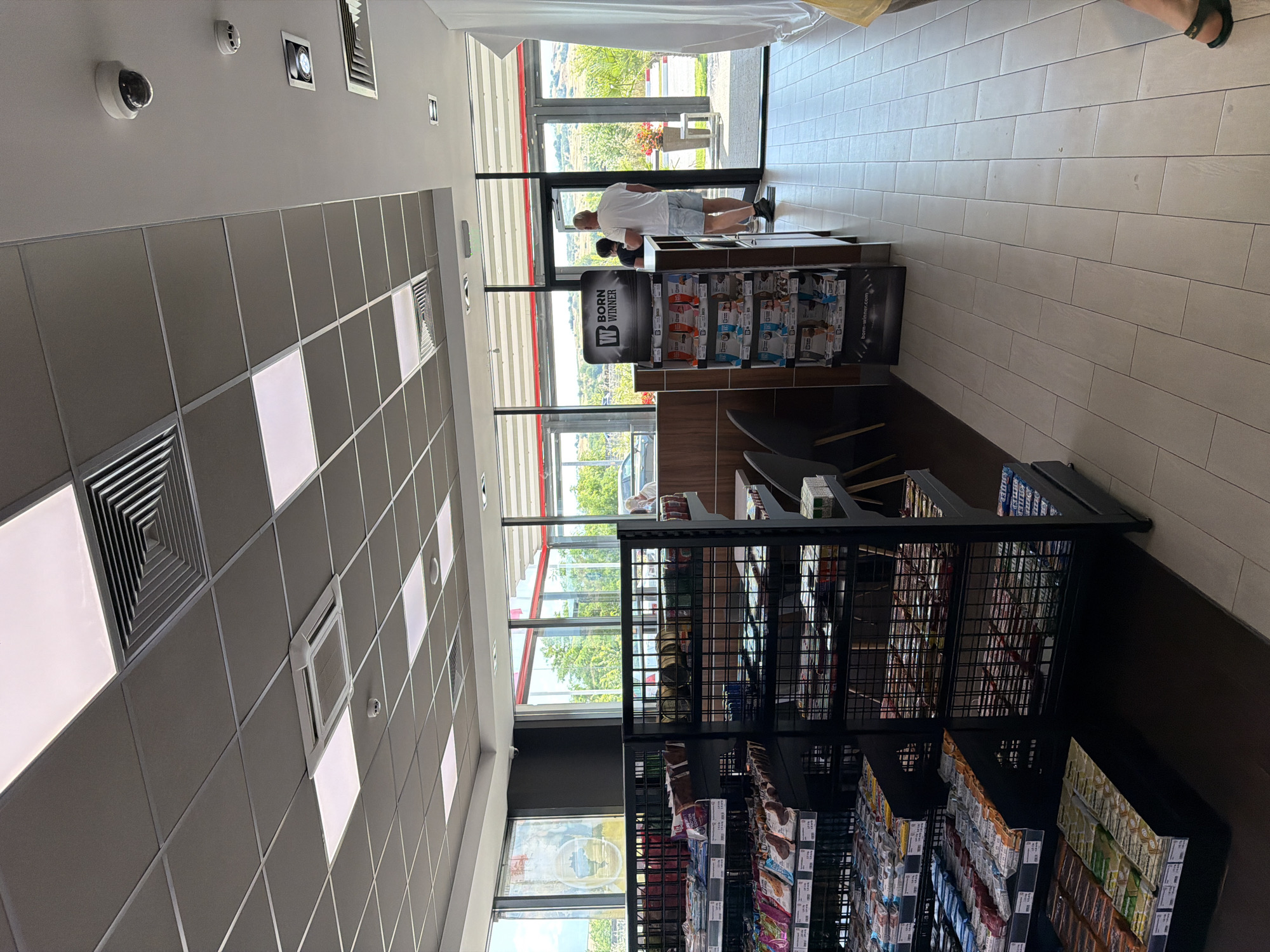 The image shows the interior of a convenience store, likely a gas station mini-mart, in Chuchuligovo, Bulgaria. The store features a drop ceiling with square white light panels and a ventilation system. Merchandise is displayed on black metal shelving units. In the foreground, shelves are stocked with various food items, including candy bars and packaged snacks. In the background, a man in a white t-shirt and blue shorts stands near the exit, looking towards the outside. Another person is partially visible behind him. The exit leads to an outdoor area with greenery and a visible road. A display stand with "BORN WINNER" branding is positioned near the entrance. The floor is tiled with light-colored rectangular tiles. A security camera is visible on the ceiling.
