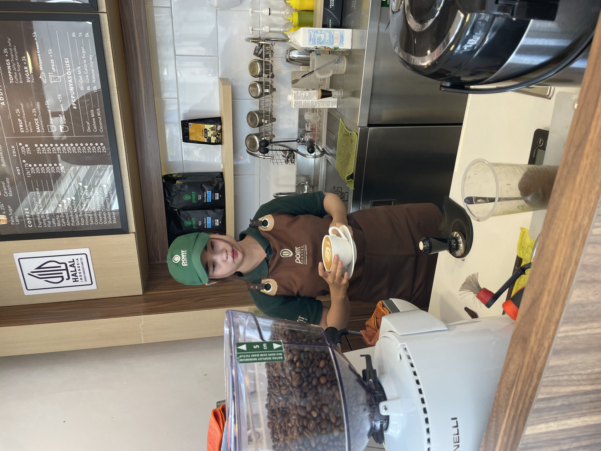 The image shows the interior of a "Point Coffee" cafe, which is likely located in Indonesia, as suggested by the "HALAL INDONESIA" logo. A barista, wearing a green cap and a brown apron with the "Point Coffee ALWAYS ON POINT" logo, is holding a cup of coffee with latte art. The cafe has a menu board listing various coffee drinks, syrups, sauces, toppings, and sugar options in Indonesian Rupiah (k). Next to the barista is a coffee bean grinder filled with coffee beans. In the background, there is a stainless steel countertop with coffee-making equipment, including coffee pumps and a dish rack. There are also bags of "Point Coffee" coffee beans visible. The scene is well-lit and appears to be a typical coffee shop setting.
