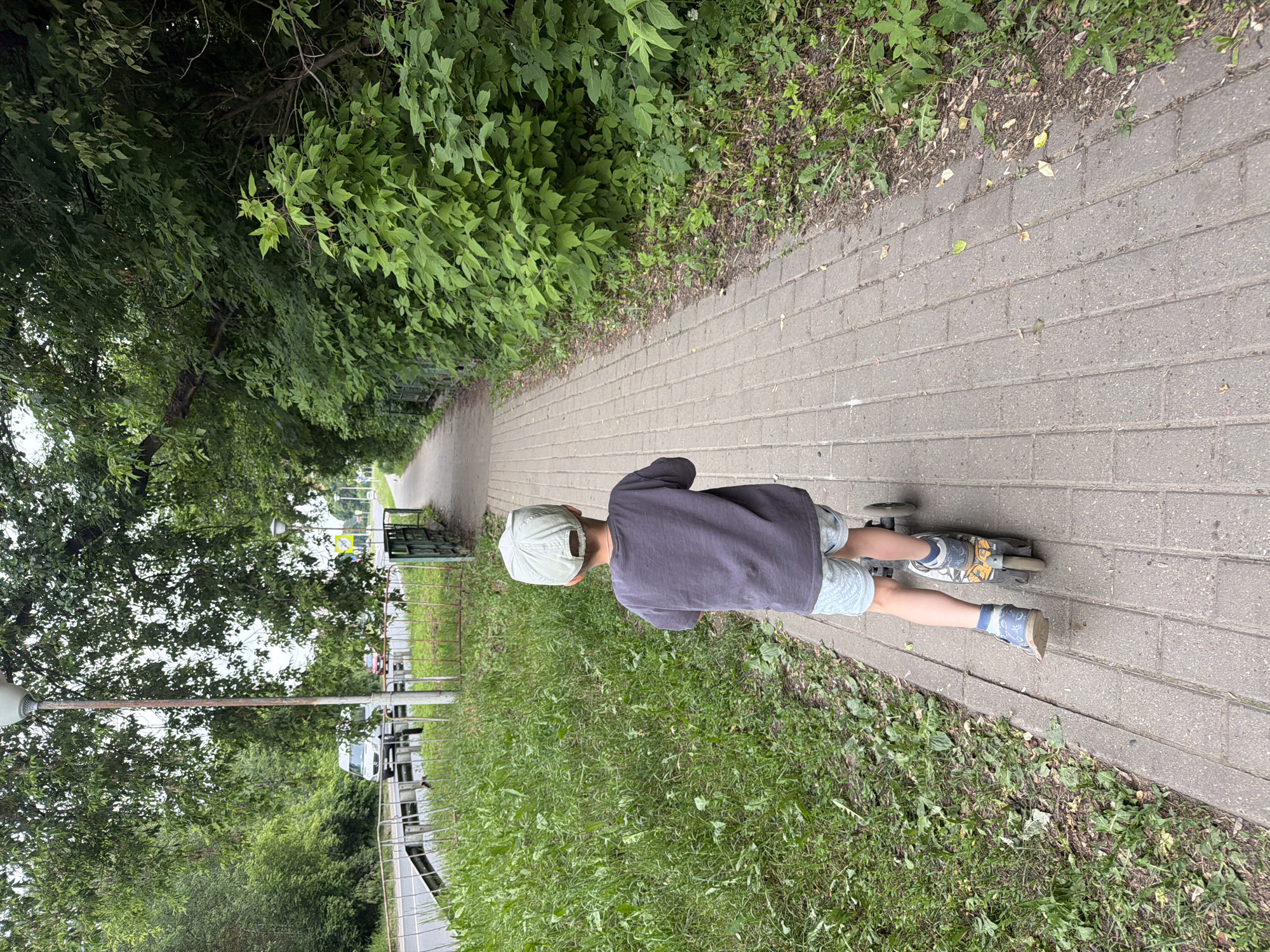 A young child wearing a grey t-shirt, light blue shorts, a light grey baseball cap, and blue and white shoes rides a three-wheeled scooter along a grey brick sidewalk. The child is facing away from the camera. To the left of the sidewalk is a grassy area with a metal fence bordering a road where a white van and two red cars are visible. Dense green foliage lines the right side of the sidewalk. A light post is visible on the left side of the image. The scene appears to be in an outdoor urban environment.