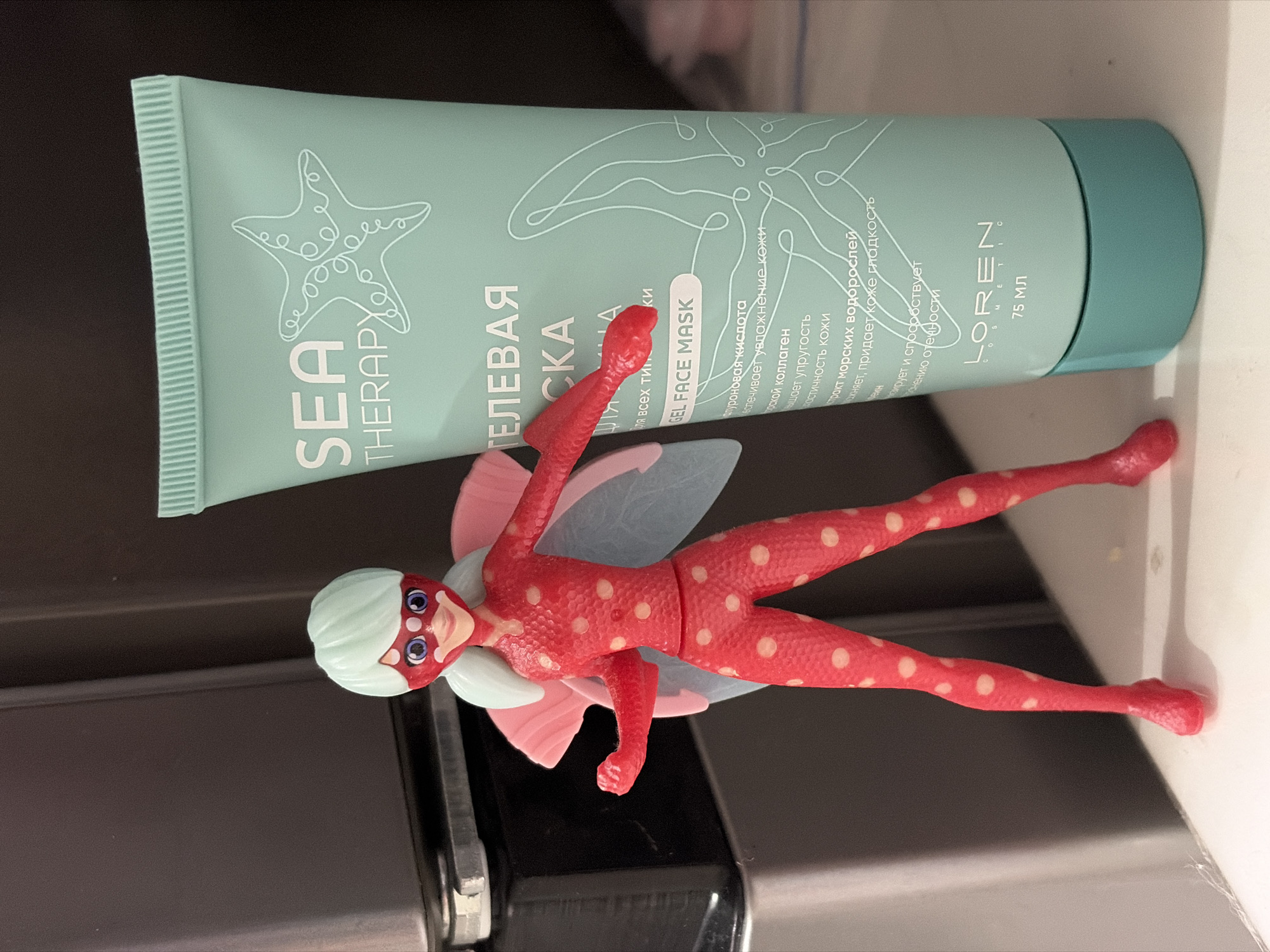 The image shows a light green tube of "Sea Therapy" gel face mask, a "Loreen" product, with Russian text and a starfish design, beside a small red polka-dotted Miraculous Ladybug action figure with a pale blue wig and wings. The background appears to be a kitchen countertop or surface with dark cabinetry. There is no indication that the image was taken in Izhevsk, Russia.