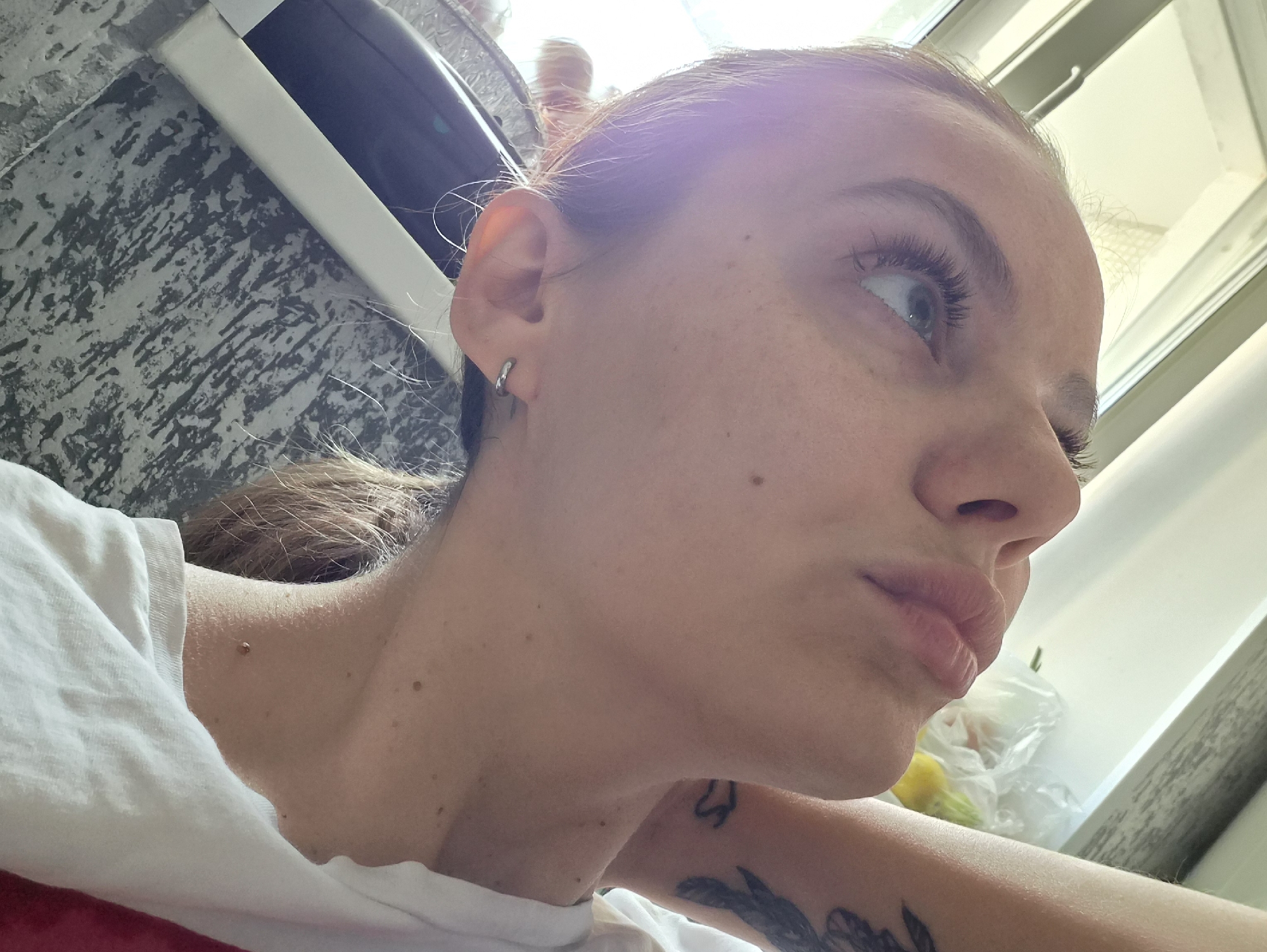 A close-up, tilted shot of a light-skinned individual with light brown hair pulled back, wearing a white t-shirt. They are looking upwards and to the right with their lips slightly pursed. Visible on their skin are freckles and a mole on the neck. An earring is visible in their left ear. Tattoos are visible on the neck and arm. The background consists of a mottled gray and white surface, a window with a white frame, and a plastic bag containing yellow and green items. No information is present within the photograph which can be used to determine if the photograph was taken in Chaikovsky, Russia.