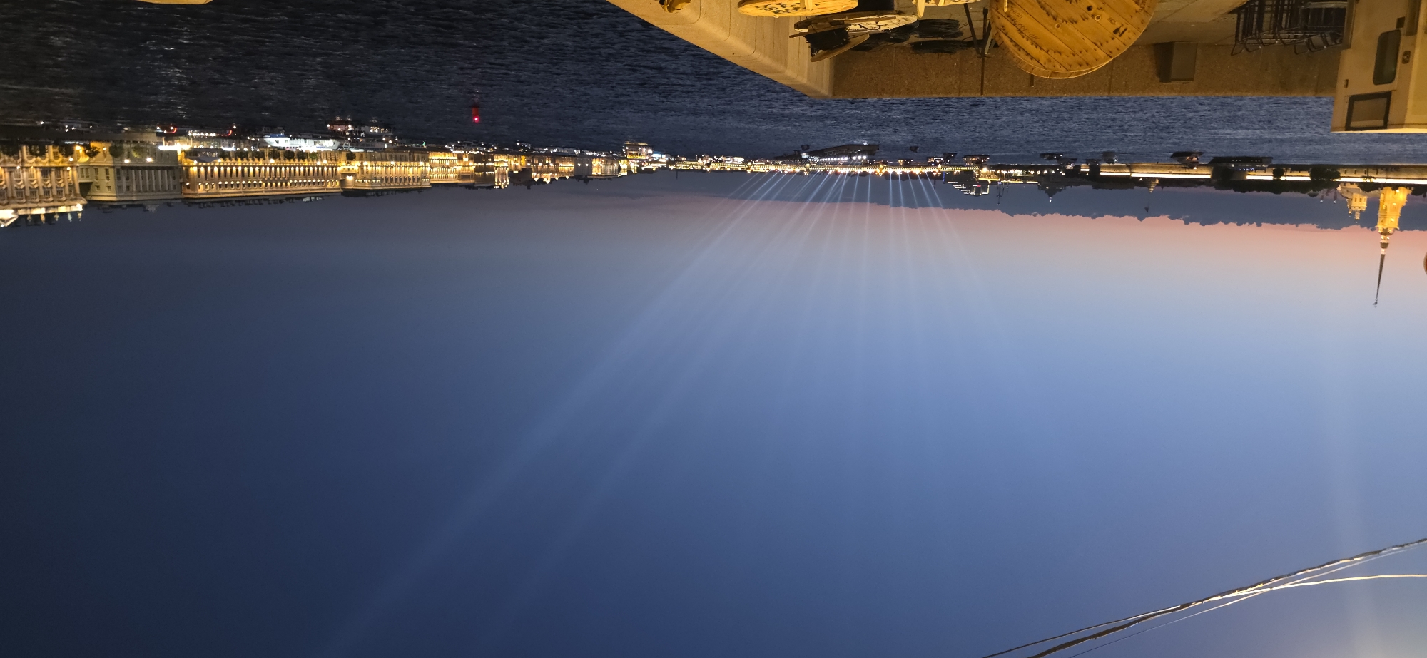 An outdoor, wide-angle, eye-level, full shot captures a twilight scene of a riverfront city. The sky is a gradient of deep blue at the top transitioning to a lighter blue with hints of pink and orange near the horizon, where scattered clouds are visible. A thin wire runs diagonally across the top of the frame.
In the foreground, the edge of a concrete embankment occupies the bottom right corner. On top of the embankment, there are several objects, including a large wooden spool, some wire, and what appears to be a piece of industrial equipment.
The river, dark and rippling, fills the middle ground of the image. A fleet of boats of various sizes are scattered across the water, some with lights illuminated.
The background features a cityscape with numerous illuminated buildings lining the opposite bank of the river. On the far left, a tall, illuminated spire with a golden top stands out against the sky, possibly part of a church or historical building. Further to the right, a long row of buildings with lit windows stretches along the waterfront. In the central background, a series of bright white light beams shoot upwards from the river towards the sky, creating a dramatic visual effect. The lighting suggests either a planned event or a natural phenomenon occurring at dusk.