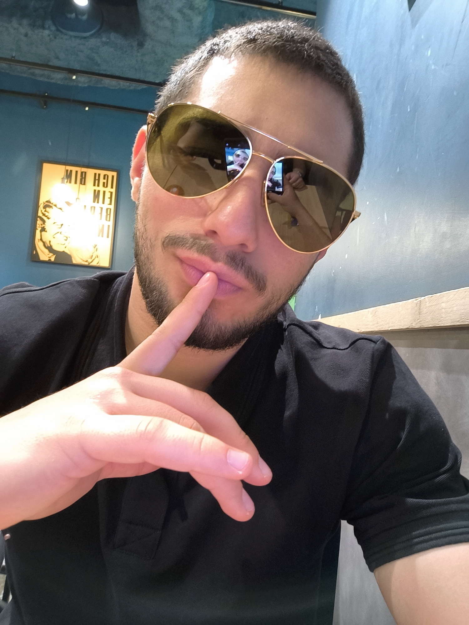 A medium close-up depicts a man with short dark hair and a beard, wearing aviator sunglasses. He is holding his index finger to his lips. He is wearing a black shirt. The background consists of a teal-colored wall with a framed black and yellow print hanging on it. The reflection in his sunglasses reveals a hand holding a phone, seemingly taking the picture. The image appears to be taken indoors. There is no information available in the image that confirms its location within the city of Strasbourg, France.