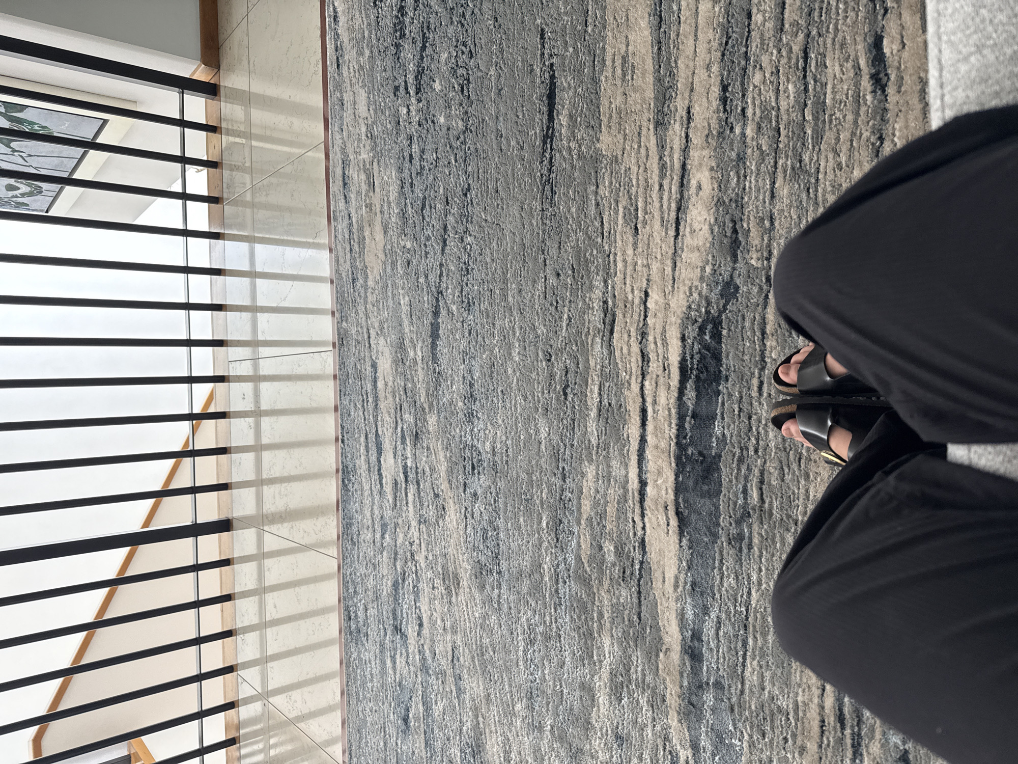 A first-person, vertically-oriented shot looking down features a person wearing black pants and black sandals with gold buckles, seated on a gray sofa. The person's feet are visible on a textured gray and beige rug with a vertical, striated pattern. To the left, a modern metal railing with horizontal black bars is visible, with light-colored tiles behind the railing. The setting is inferred as an indoor space with natural light entering from the left. There is no available information on San Bernardino Tlaxcalancingo, Mexico.