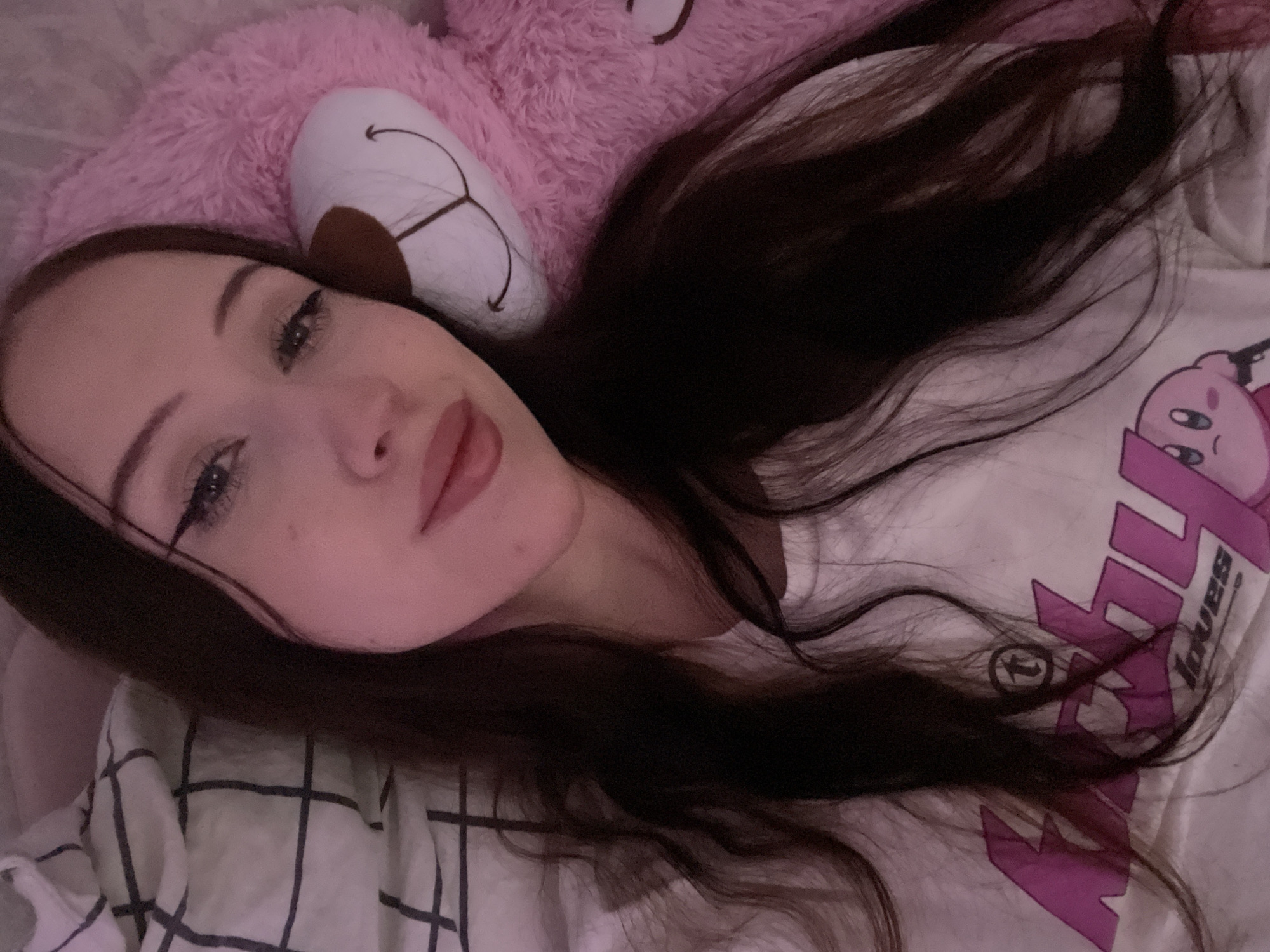 A young woman with long, dark hair lies on a pink, fuzzy object, possibly a large stuffed animal, and a white checkered blanket. She is wearing a white t-shirt with a graphic print of a pink character, likely Kirby, on it. Her eyes are open and she is looking towards the camera with a soft expression. Her makeup includes winged eyeliner and lip gloss. The overall mood of the image is relaxed and intimate. The lighting suggests it might be nighttime or a dimly lit room, and the pink hue from the stuffed animal casts a soft glow on her skin. No specific location cues are visible beyond what's described.