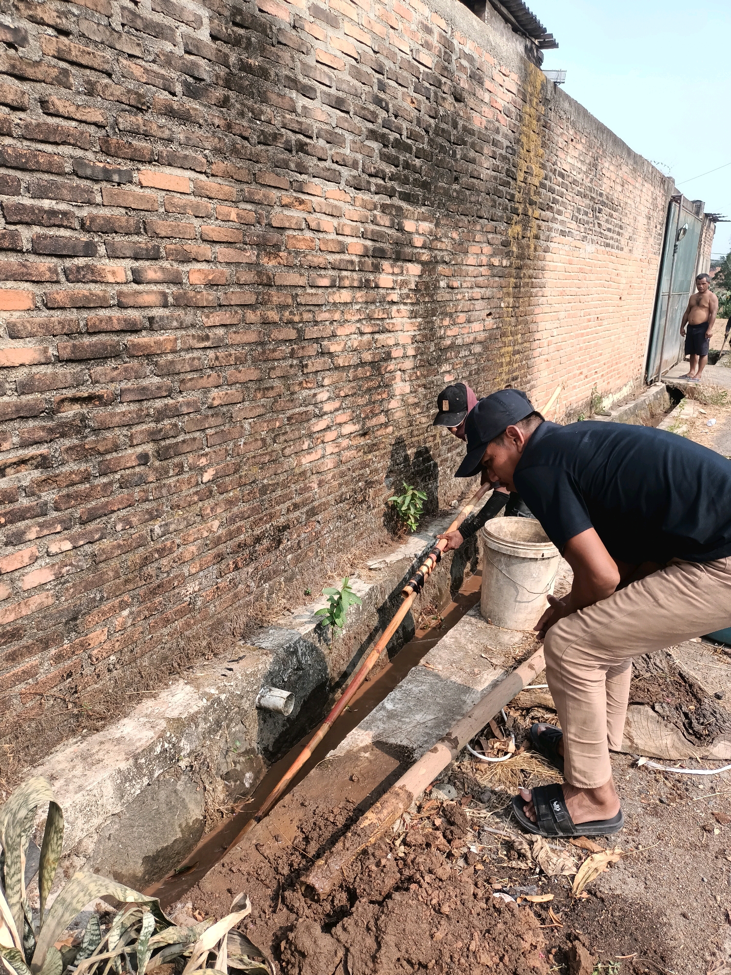 In Metro, Indonesia, two men are working on clearing a muddy drainage ditch alongside a brick wall. The man in the foreground, wearing a black t-shirt and tan pants, is leaning down, with his hands near the edge of the ditch. A second man, also wearing a black t-shirt and a black baseball cap, is further up the ditch, holding a long wooden pole that is inserted into the muddy water. A white bucket sits on the ground next to the man in the foreground. The ditch appears to be filled with mud and debris. In the background, a man without a shirt is standing near a large metal gate. The environment is outdoors, with a clear sky.