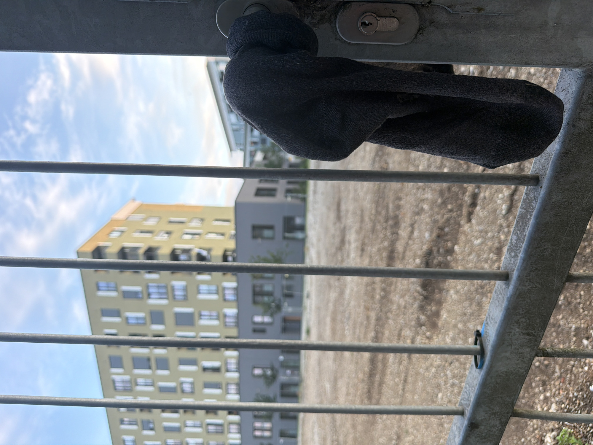 The image depicts a close-up view of a metal gate or fence in an urban outdoor setting. A dark-colored sock is hanging from the handle of the gate. The background features modern buildings, one of which is a multi-story yellow building with numerous windows. Another lower building with a darker facade is also visible. The ground appears to be unpaved and dusty, possibly a construction site or an undeveloped area. The sky above is partly cloudy, with visible sunlight. The composition suggests a candid shot, with the focus on the sock and the gate. The overall impression is one of an everyday, perhaps slightly unkempt, urban environment. The location is not explicitly indicated in the image, but if it were in Munich, Germany, it would likely be in a residential or mixed-use area undergoing development or renovation.