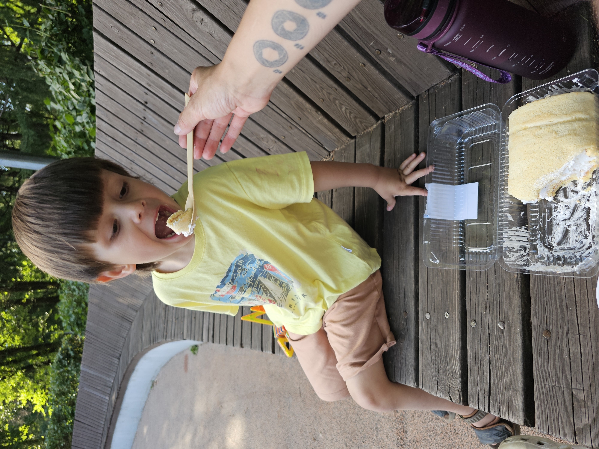 In this outdoor scene, a young child wearing a yellow t-shirt and tan shorts is being fed a piece of cake from a wooden spoon. The child's mouth is open, ready to receive the treat. The child is seated on a wooden bench or platform, which is part of a larger wooden structure. To the right of the child, there are two clear plastic containers, one empty and one holding a slice of cake. A large, dark purple water bottle with a purple strap rests on the wooden surface. The background shows lush green trees and foliage, suggesting a park or natural setting. The sunlight filtering through the leaves indicates it's daytime. A person's arm with a tattoo of concentric circles is visible in the upper part of the image, holding the spoon. The wooden planks have visible nails and grain patterns, contributing to a rustic aesthetic. The overall mood appears relaxed and pleasant, possibly a family outing or picnic.