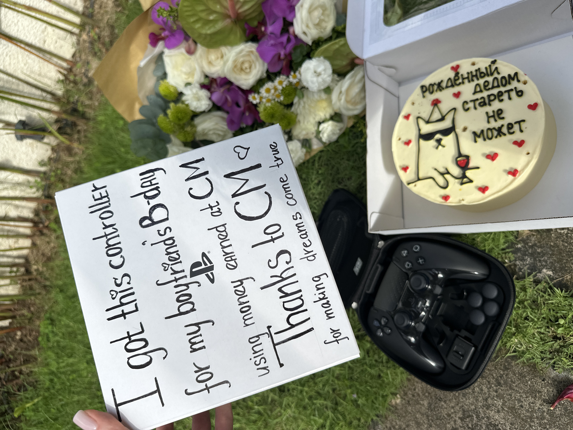 A gift layout on an outdoor surface, presumably in North Kuta, Indonesia, includes a bouquet of white roses and purple orchids wrapped in brown paper, a white cake with a cartoon dog wearing sunglasses and a party hat, and a black PlayStation controller in a black zip-up case. A white sign with handwritten black ink states, "I got this controller for my boyfriend's B-day, using money earned at CM. Thank's to CM for making dreams come true." The cake has Cyrillic text that reads, "рожденный дедом стареть не может." The surface appears to be a combination of grass and concrete. A hand with a manicured nail is holding the sign.