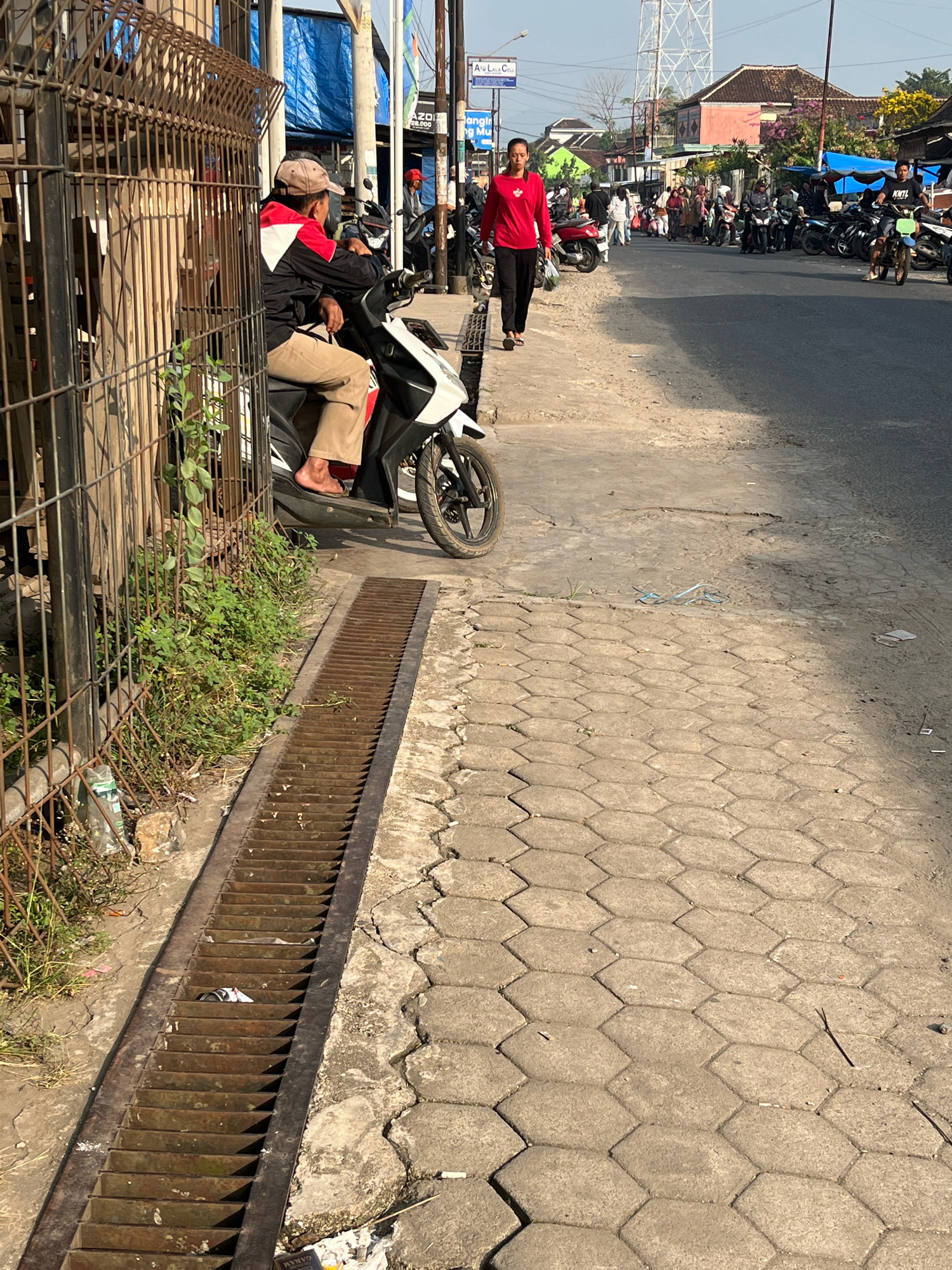 A medium shot captures a street scene in Kemuning, Indonesia, on a sunny day. In the foreground, a rusty metal grate runs along the edge of a hexagonal-tiled sidewalk. To the left, a person sits on a white and black motorbike, wearing a beige cap, a black and red jacket, and beige pants. Their bare feet are visible. The person appears to be looking to their right. Behind the motorbike, a wire fence borders a patch of green foliage. In the background, a woman in a red long-sleeved shirt and black pants walks on the sidewalk, carrying a dark bag in her left hand. Motorbikes are parked along the street on the right side, and several people are visible further down the road, some on motorbikes and others walking. Buildings with various colored roofs line the street. A tall white tower stands in the distance. The environment appears to be a bustling urban area with clear skies.