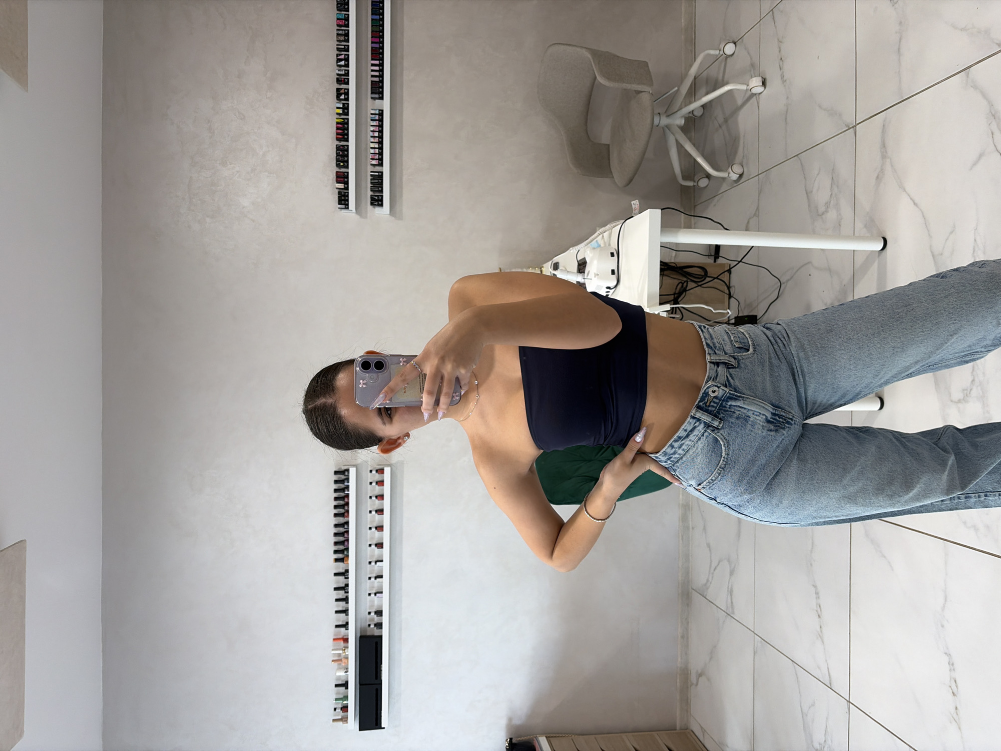 The image depicts a female subject taking a mirror selfie inside a commercial establishment, specifically a nail salon, located in Sofia, Bulgaria. The subject, visible from the mid-thigh up, has dark hair pulled back and wears a dark blue strapless crop top, light blue wide-leg jeans, a thin silver bracelet on her left wrist, and a delicate necklace. She holds a light purple-cased smartphone with her right hand, obscuring part of her face, while her left hand rests on her hip.
The interior features a light grey textured wall, a white tiled floor with a marble effect, and a white ceiling with a rectangular panel. On the left, partially visible, are a light wooden drawer unit and a dark green upholstered chair. Two white shelves are mounted on the left wall, displaying rows of small bottles, presumably nail polish, alongside two black rectangular items on the top shelf. To the right of the subject, a white rectangular table holds a white electronic nail drill/file machine, another smaller white device, and black cables. Adjacent to the table is a light beige upholstered office chair with a white five-star base and wheels. Two additional white shelves, similar to those on the left, are mounted on the right wall, also displaying numerous small bottles of nail polish. The overall environment appears clean, modern, and well-lit.
