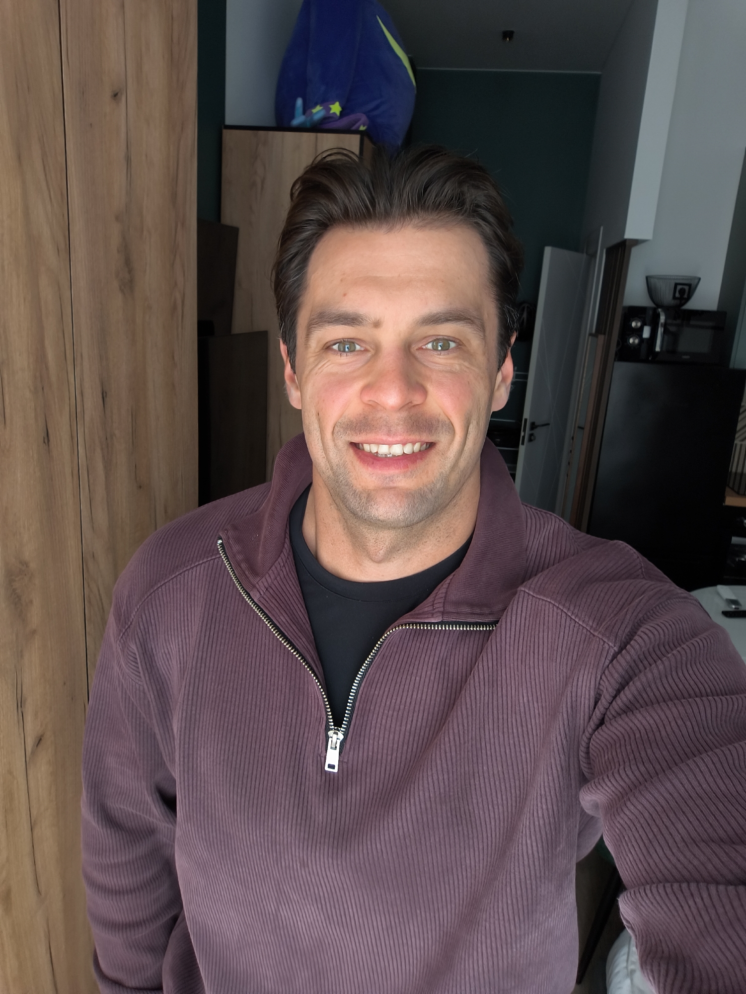 A man, appearing to be in his late 30s, is captured in a selfie taken from a medium close-up perspective. He has dark brown hair styled casually and striking green eyes. A warm smile plays on his lips, revealing white teeth. He is wearing a deep purple, ribbed, quarter-zip sweatshirt over a black t-shirt.
The background suggests an indoor setting, possibly an apartment. To the left, a wooden wardrobe or cabinet with vertical paneling dominates the frame. Behind him, on a shelf, a plush blue toy with yellow accents is visible, and further back, a dark teal painted wall can be seen. To the right, a black refrigerator and other kitchen appliances are present, along with a white door. The overall impression is that of a casual, personal space. This scene is unlikely to be in Moscow, Russia based on the visual cues.