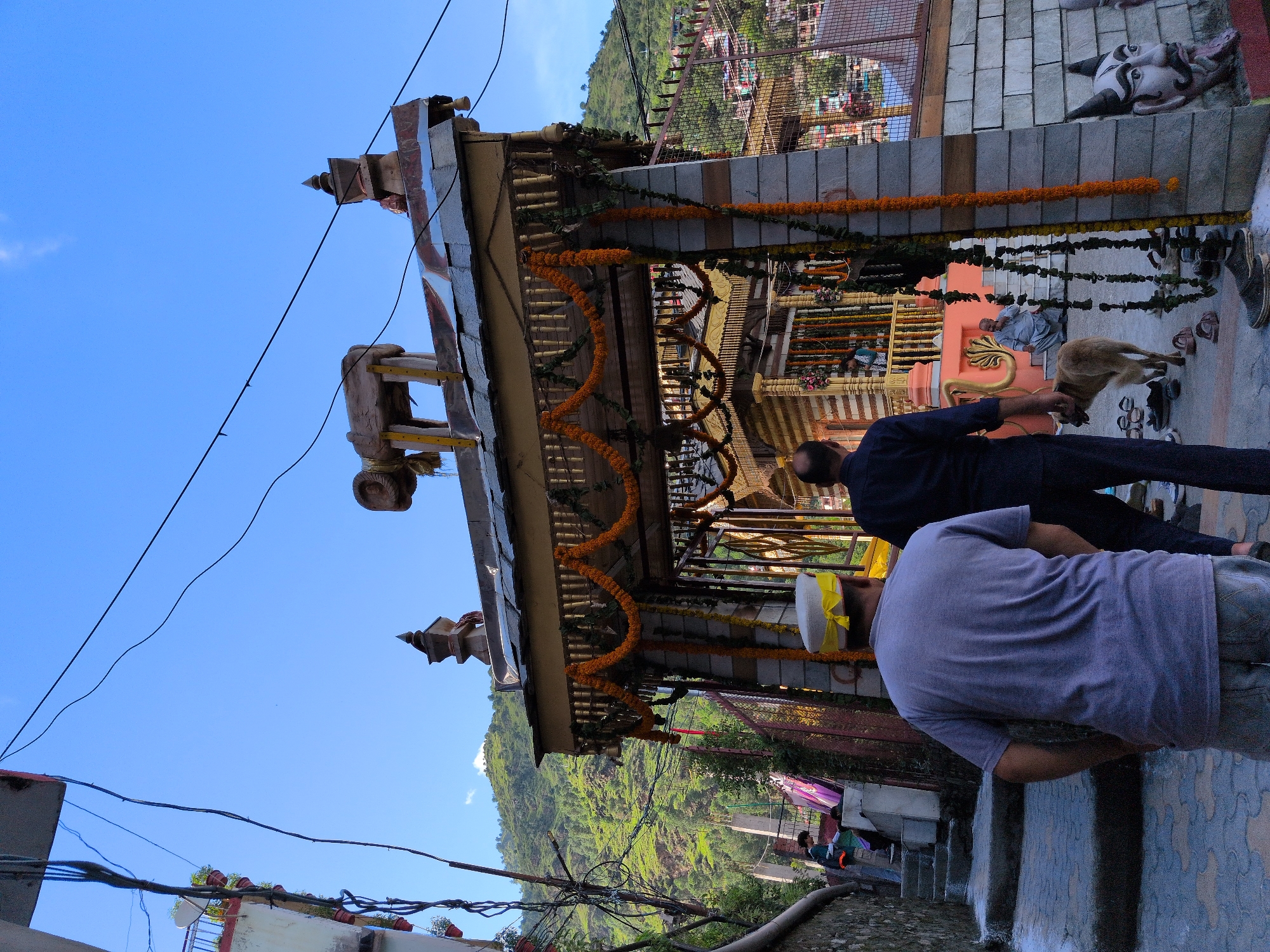 The image depicts an outdoor scene in the city of Kullu, India, likely near a temple or religious site. In the foreground, two men are walking away from the camera, one wearing a grey t-shirt and a traditional Himachali cap with a yellow band, and the other in a dark blue shirt. A light brown dog is walking ahead of them. Behind the men, an ornate entrance structure, possibly a gateway to a temple complex, is visible. This structure is decorated with garlands of orange marigolds and green foliage. Beyond the entrance, the interior of a building, likely the temple, can be seen with intricate carvings and decorations. A person is seated on the steps inside the temple complex. In the background, a lush green mountainside with scattered buildings is visible under a clear blue sky with scattered clouds. The scene is characterized by a mix of traditional architecture and natural beauty, common in the Kullu Valley. The presence of the dog and the casual attire of some individuals suggest a public area, possibly a tourist or pilgrimage site. Electrical wires are strung overhead, indicating the presence of modern infrastructure. A carved architectural element resembling a face with horns and a protruding tongue is visible on the right side of the image, on the wall of a building.