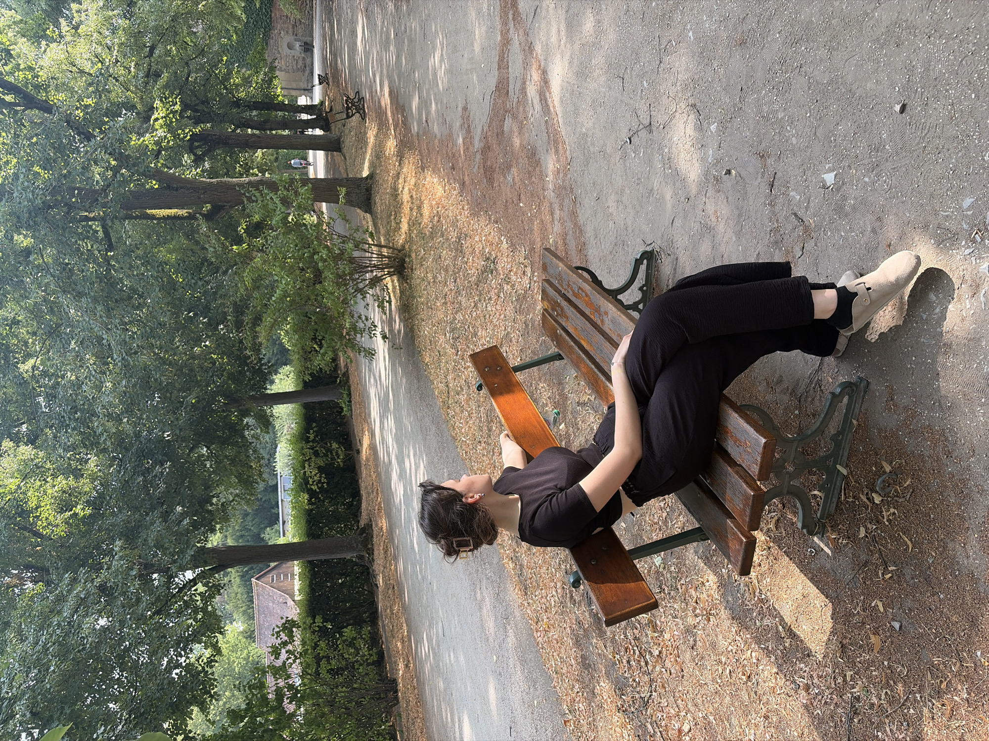 A person is sitting on a wooden park bench in the Hofgarten, Würzburg, Germany. The person is wearing a black top and black pants, with light-colored shoes. They are looking to the right, away from the camera. The bench is old with a dark green metal base. The ground around the bench is covered in dirt and fallen leaves. In the background, there are trees, a path, and other park benches. The sun is shining, casting shadows on the ground. A building with a brown roof is visible in the distance.