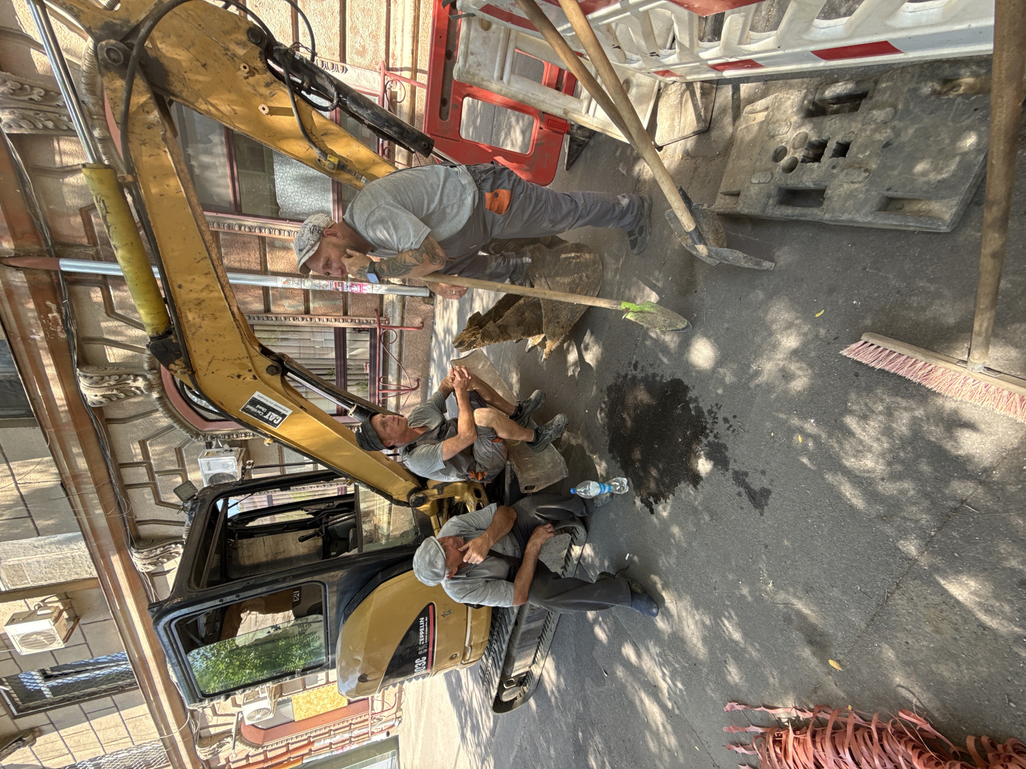 Three construction workers are taking a break next to a yellow excavator on a sunny day in an urban environment. The excavator is parked on a paved surface, likely a street or sidewalk, with buildings visible in the background. One worker, wearing a gray shirt and pants with a grey flat cap, is seated on the excavator's track, resting his chin on his hand and looking down. Another worker, in a gray t-shirt and dark shorts with an orange pocket detail, is sitting on the excavator's bucket, also with his chin in his hand, looking to the side. The third worker, in a gray t-shirt and grey pants, is standing near the excavator, leaning on a shovel, and looking to the left, with his hand on his chin. A bottle of water and a cigarette butt are on the ground near the seated workers. Various construction tools, including shovels and what appears to be a large concrete block, are scattered around the scene. Red plastic fencing and a red rope or hose are also visible. The environment appears to be dusty and sun-drenched, with shadows cast by overhead trees.