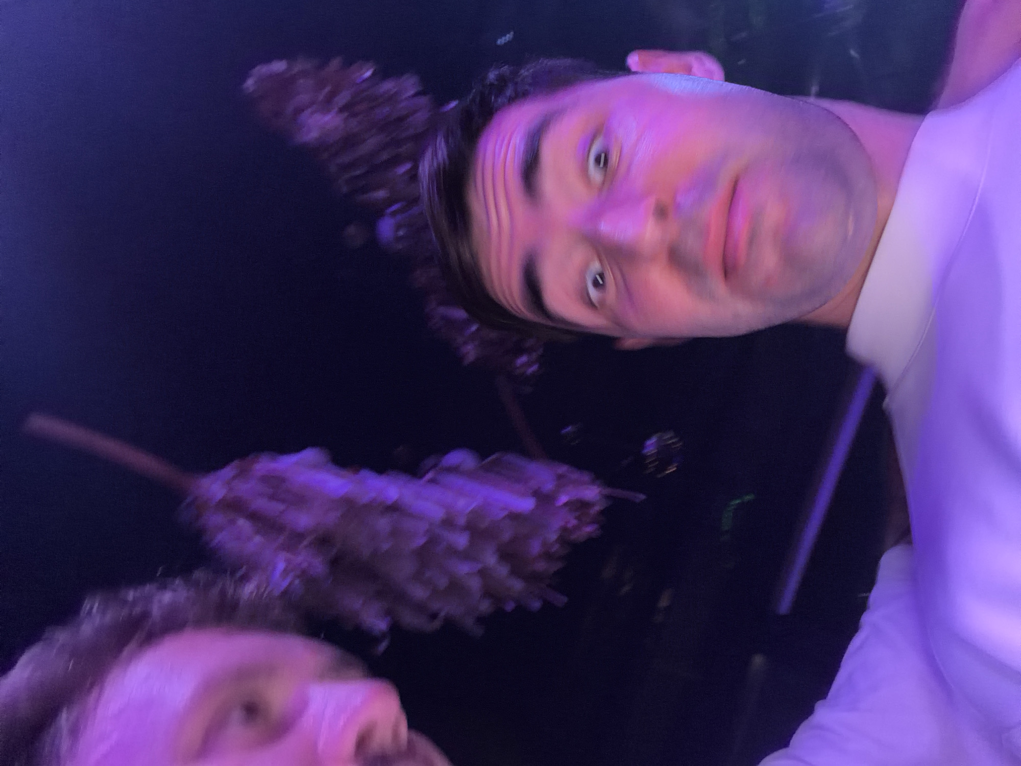 A slightly blurred selfie taken in a dark environment with a purple light source. A man with short dark hair and light skin is in focus. He is wearing a white collared shirt. Another person is partially visible in the bottom left corner. In the background, there appears to be a blurred, indistinct object, possibly a decorative piece, and a dark background. The specific location within Choeng Thale, Thailand, is not discernible from the image.
