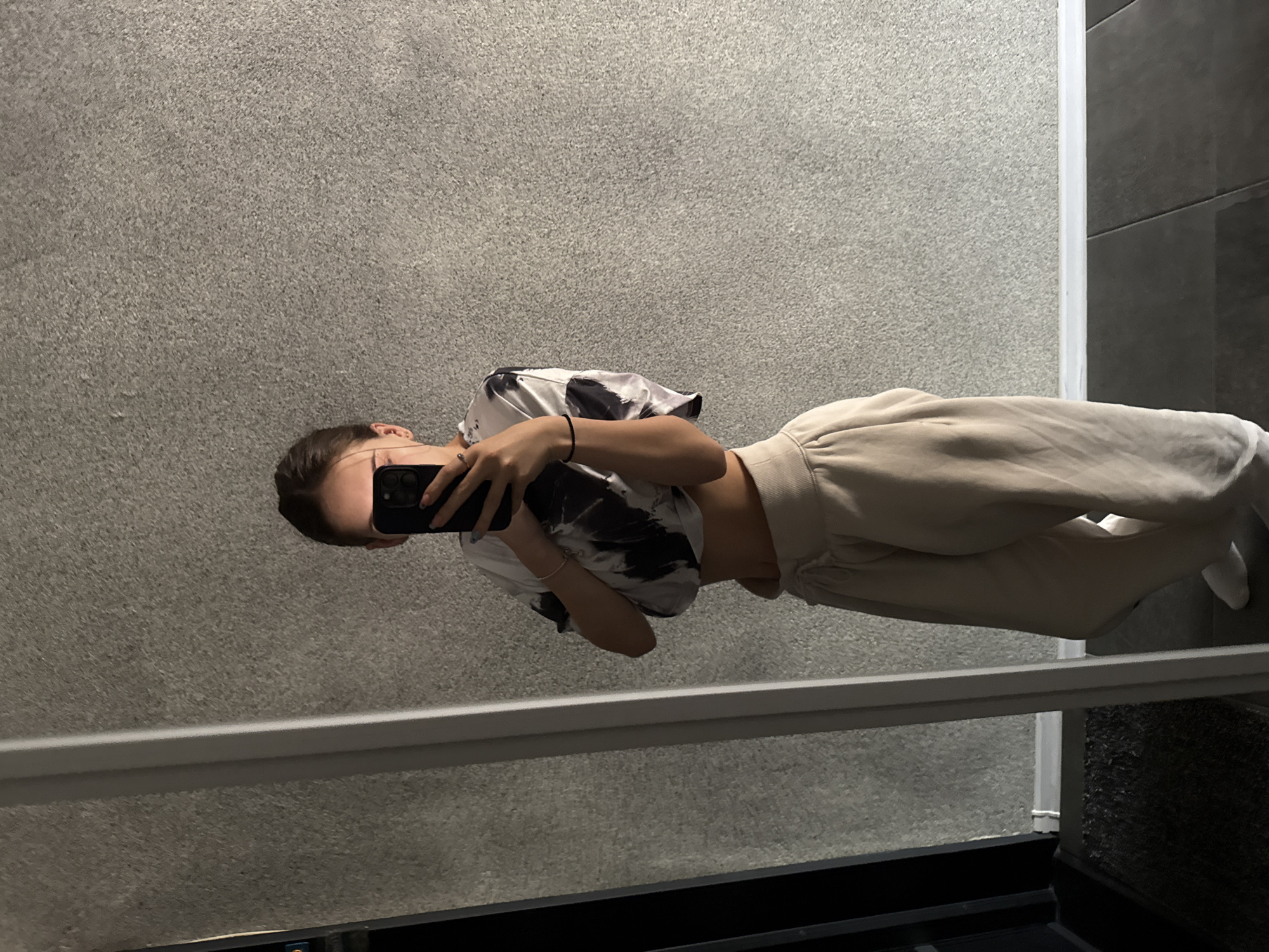 A rotated image shows a person taking a mirror selfie in an indoor setting, likely in Sofia, Bulgaria. The individual's face is obscured by a black smartphone held up to capture their reflection. They are dressed in a black and white tie-dye or patterned crop top, revealing a midriff, and light-colored, possibly beige or cream, wide-leg trousers. A dark bracelet is visible on one wrist, and rings adorn the fingers holding the phone.
The background features a textured, speckled grey wall behind the person, meeting a darker grey or black tiled wall on the right, separated by a distinct white vertical strip. A silver-grey horizontal rail or ledge runs across the lower portion of the image, reflecting the environment. The smartphone itself is prominent, showcasing its multi-lens camera system. The overall lighting suggests an interior space with artificial illumination. No visible text is present in the image.