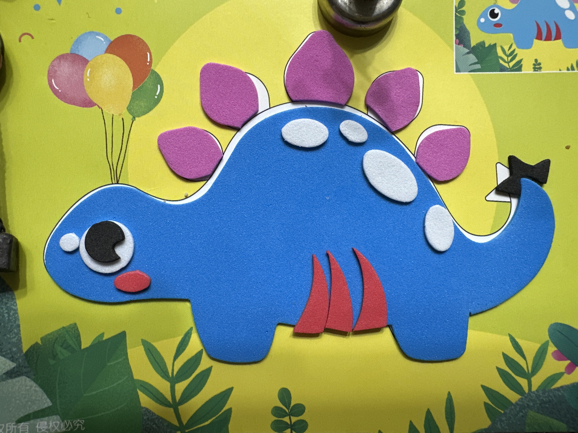 The image shows a colorful, cartoonish representation of a dinosaur, likely made of foam or similar soft material, adhered to a yellow background. The dinosaur is blue with pink dorsal plates, white spots, red belly plates, and a black bow on its tail. It has a large, cartoon eye with a white and black pupil and a small red nostril. To the left of the dinosaur, there's a cluster of balloons in various colors. The background is decorated with green foliage. A sticker of a similar dinosaur is placed in the upper right corner. The image seems to be taken indoors, with some metal hardware visible in the upper center and left corner. No specific location in Baranovka, Russia, can be determined from the image.