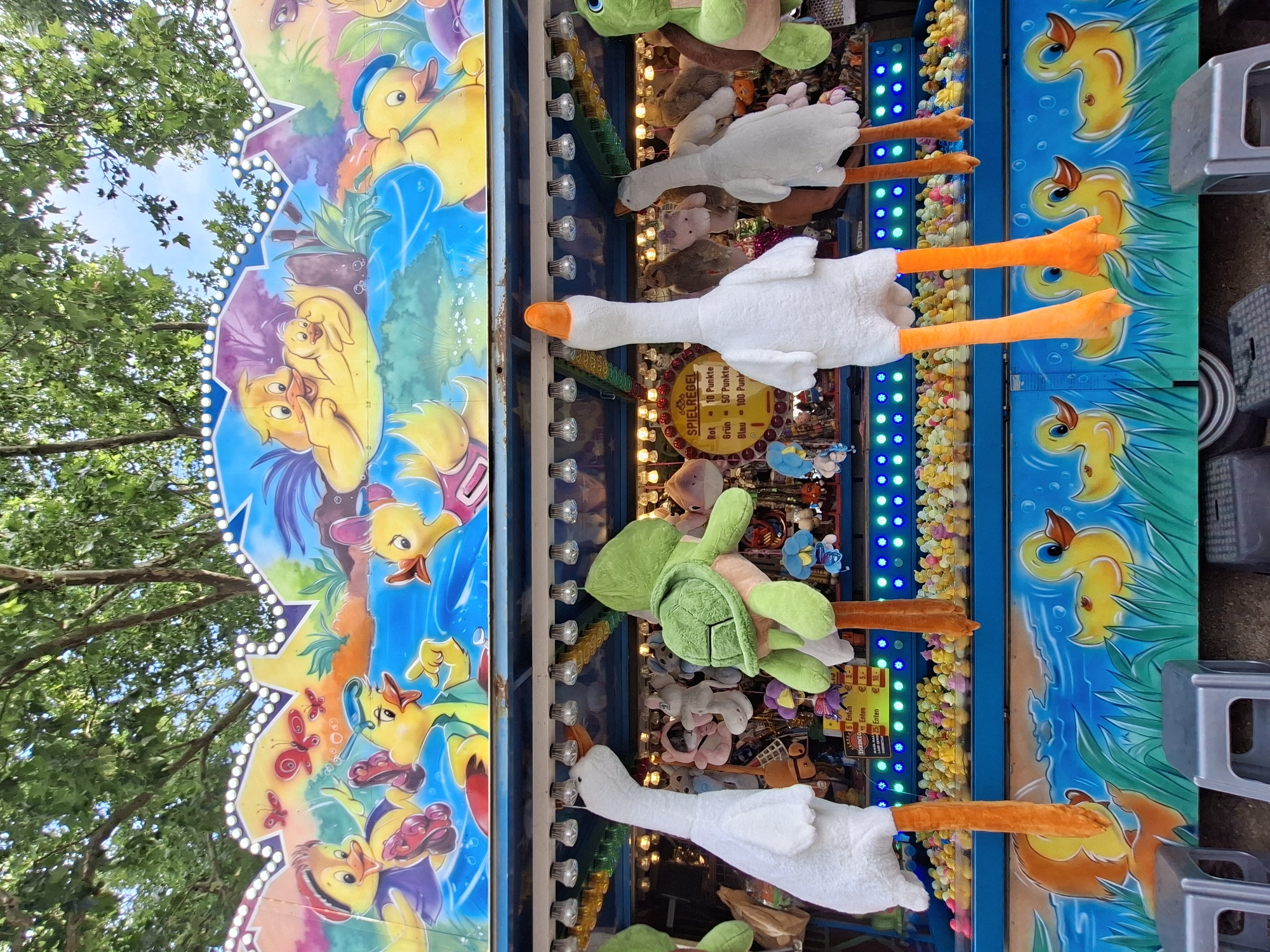 This image depicts a carnival game stall at the "Volksfest" in Wiesbaden, Germany. The stall features a colorful mural of cartoon ducks and butterflies across the top. Below the mural, a row of plush toys, including geese and a turtle, hang suspended from the stall. The main attraction of the game appears to be a pool of small plastic ducks, with a sign displaying game rules and prices. The stall is set up outdoors, with trees visible in the background and what appear to be metal stools in the foreground. The overall atmosphere suggests a festive event.