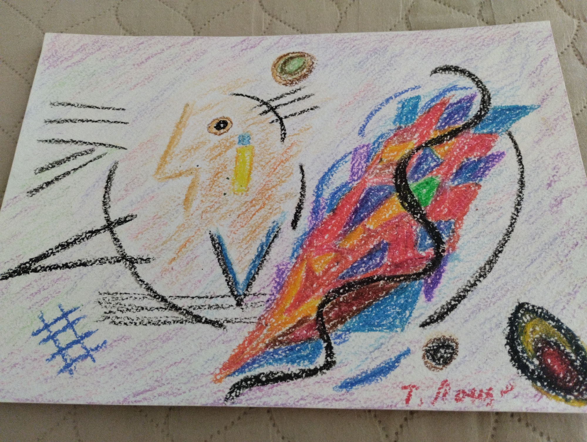 The image is an abstract drawing on white paper, likely created with crayons or colored pencils, laid on a patterned textile surface. The drawing features a stylized face-like form in orange on the left, with a yellow rectangular shape and an eye indicated by a small brown circle. Several black lines and shapes are scattered across the drawing, including parallel lines on the left and a curved line dominating the center. A vibrant, fragmented shape in the center is composed of red, blue, purple, and yellow segments, suggesting a dynamic or energetic element. There are also smaller circular and oval shapes, one with concentric rings of color, on the right side. The background of the drawing is lightly filled with purple and green pastel strokes. A signature, "T. Nous," is visible in red at the bottom right. The scene does not depict any specific location in Mezitli, Turkey.