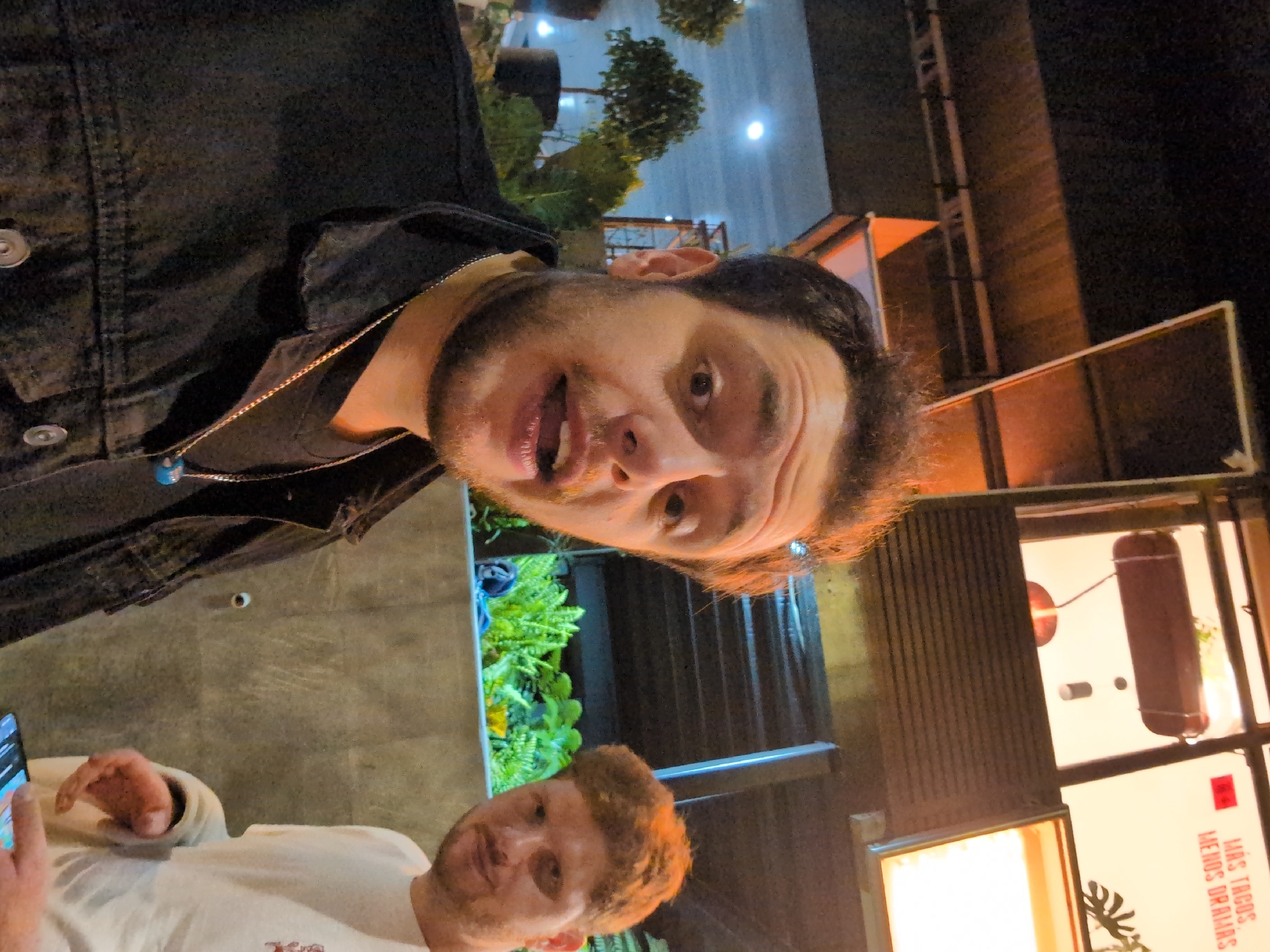 Two men are in an outdoor location with food establishments in Mexico City, Mexico. One man is wearing a black jacket and a necklace. The second man is wearing a light-colored shirt. The men are likely interacting with each other. The background features plants, streetlights, and buildings.