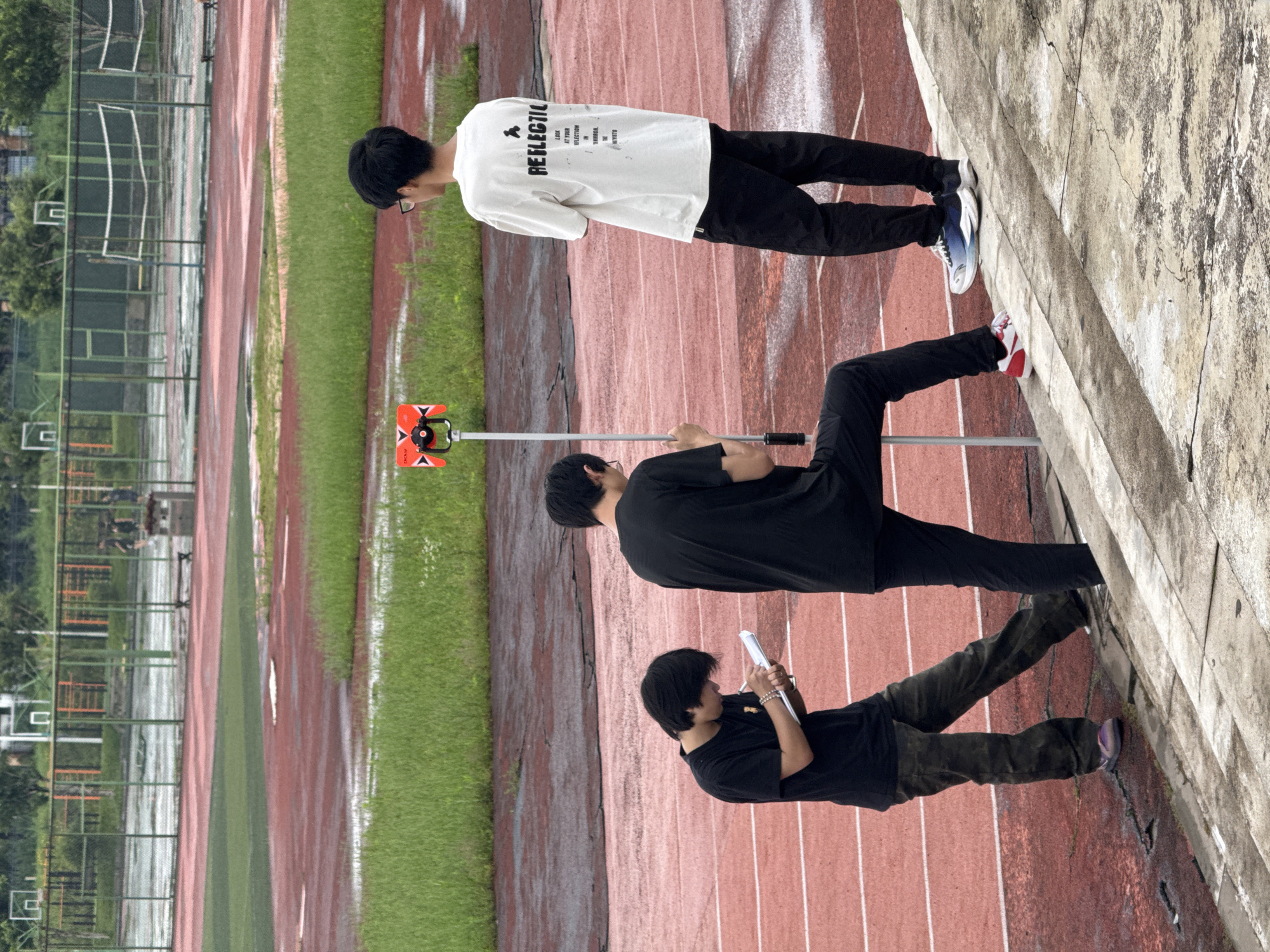 The image depicts three young males on a wet, red running track with green grass and a fence in the background. In the foreground, one male in a black t-shirt and camouflage pants is writing on a notepad. To his right, a second male in a black t-shirt stands with one foot on a concrete step, looking towards the right. Further to the right, a third male in a white t-shirt with text and black pants faces away from the camera, looking towards the right. A surveying prism is mounted on a pole between the first two males. The scene appears to be a sports facility, possibly within the city of Jiangjin, China, given the context provided. The sky is overcast, contributing to the wet appearance of the track and surrounding grass.