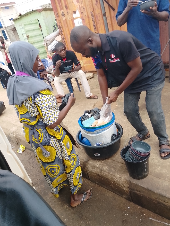 A street scene shows a man in a black "HairSense" shirt handing a plastic bag to a woman in a yellow patterned dress and gray hijab. They stand next to a large black tub filled with a blue and white bucket, plastic plates, and other items. Another man sits on a stool in the background eating from a bowl. A pile of black plastic bowls sits in a black bucket. A shipping container and various buildings are visible in the background.
