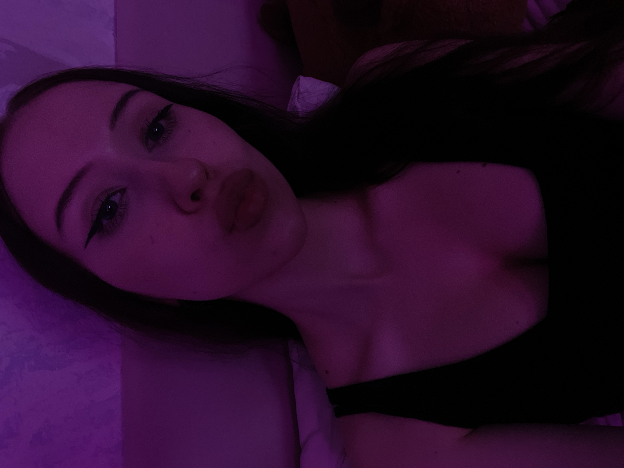 A young woman with long dark hair and prominent eyeliner poses for a selfie. She is wearing a black tank top. The lighting in the image is predominantly purple, casting a pinkish hue on her skin and the textured wall behind her. The image appears to be taken indoors, likely in a bedroom given the presence of what looks like a pillow. There is no indication of a specific location within Votkinsk, Russia.