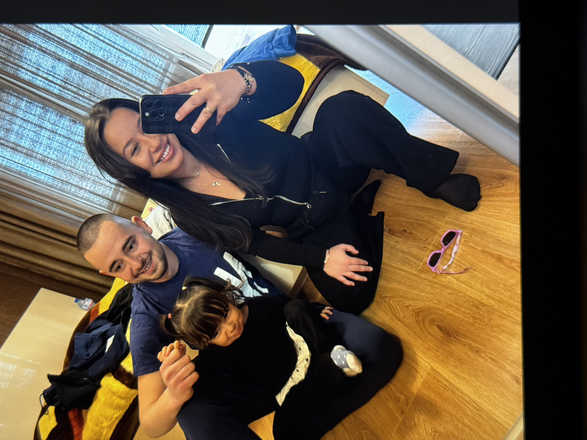 The image depicts a woman taking a selfie with a mobile phone, a man, and a young girl. They are located indoors, sitting on a wooden floor. The woman is wearing a black zip-up jacket and black pants. The man is wearing a blue t-shirt. The young girl is wearing a black long-sleeved shirt and navy pants. Next to them on the floor are pink sunglasses. Behind the group is a chair with a yellow and brown blanket. Location is Unknown, Unknown.