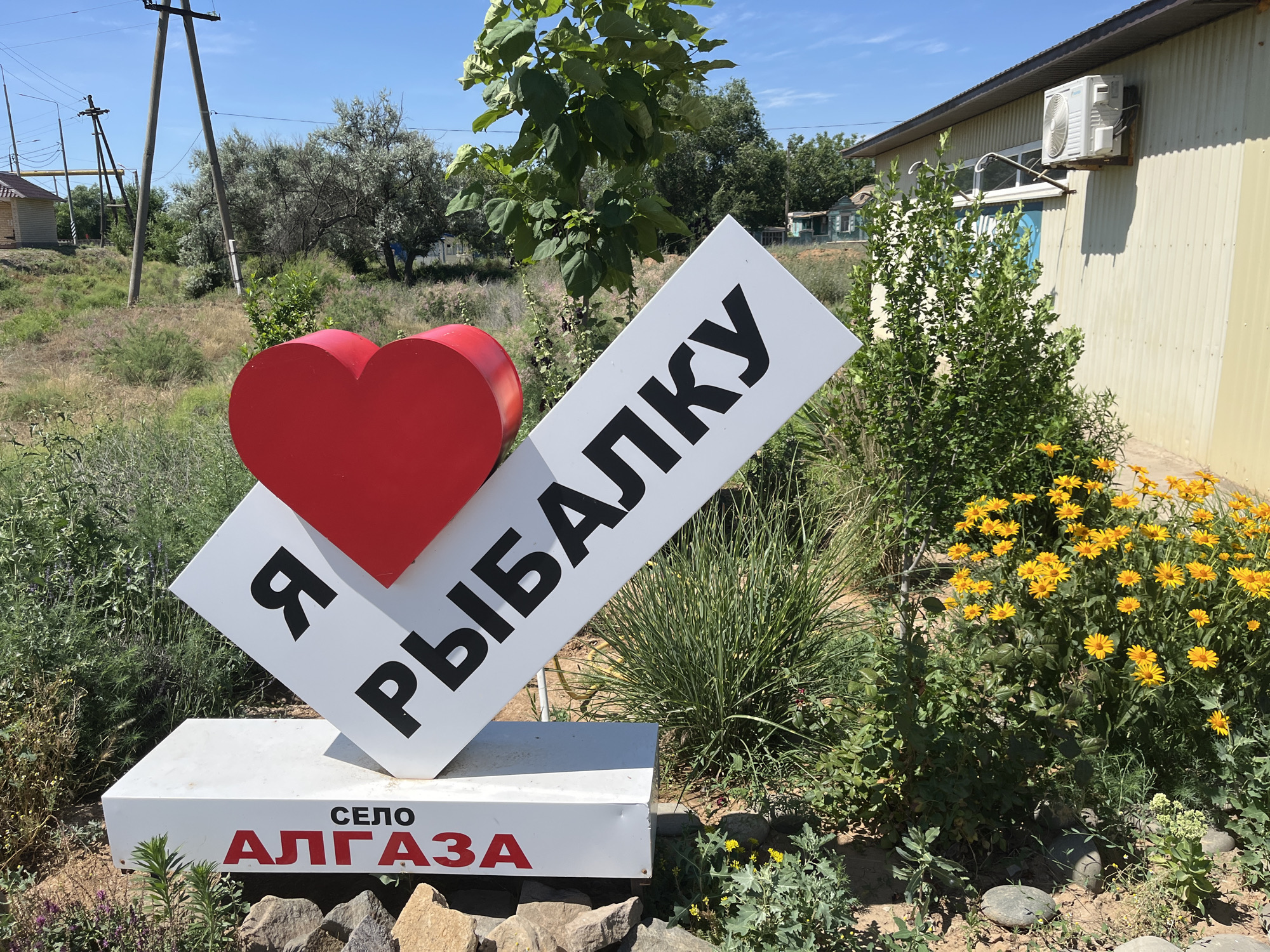 The image captures a decorative sign in the village of Algaza, Russia. The sign features a large red heart on the left side, mounted on a white, angled surface that displays Russian text in black capital letters: "Я РЫБАЛКУ". Below this, a white rectangular base has the words "СЕЛО АЛГАЗА" printed in black and red capital letters respectively. The sign is positioned outdoors in a sunny environment with a clear blue sky. To the right of the sign, a cluster of bright yellow flowers adds a splash of color. In the background, various types of vegetation, including trees and bushes, are visible, along with a building that appears to be made of corrugated metal and a small house in the distance. Electrical poles and wires are also present in the scene.