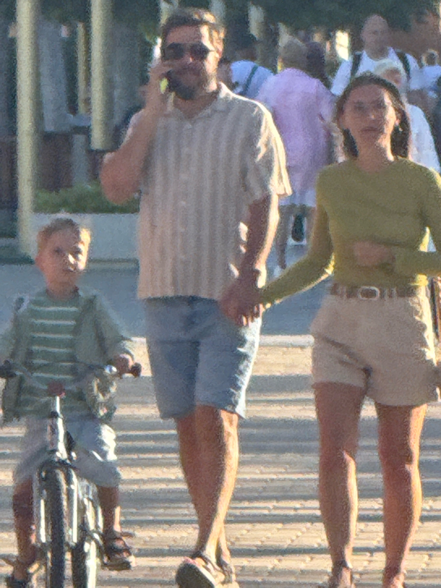 In Anapa, Russia, a man, a woman, and a child are walking on a boardwalk. The man, on the left, is wearing sunglasses, a striped shirt, and denim shorts. He is holding his phone to his ear. The woman, on the right, is wearing a green long-sleeved shirt and beige shorts, and is holding the man's hand. The child, in the foreground, is riding a bicycle and wearing a striped shirt. The environment appears to be a sunny outdoor area with other people in the background.
