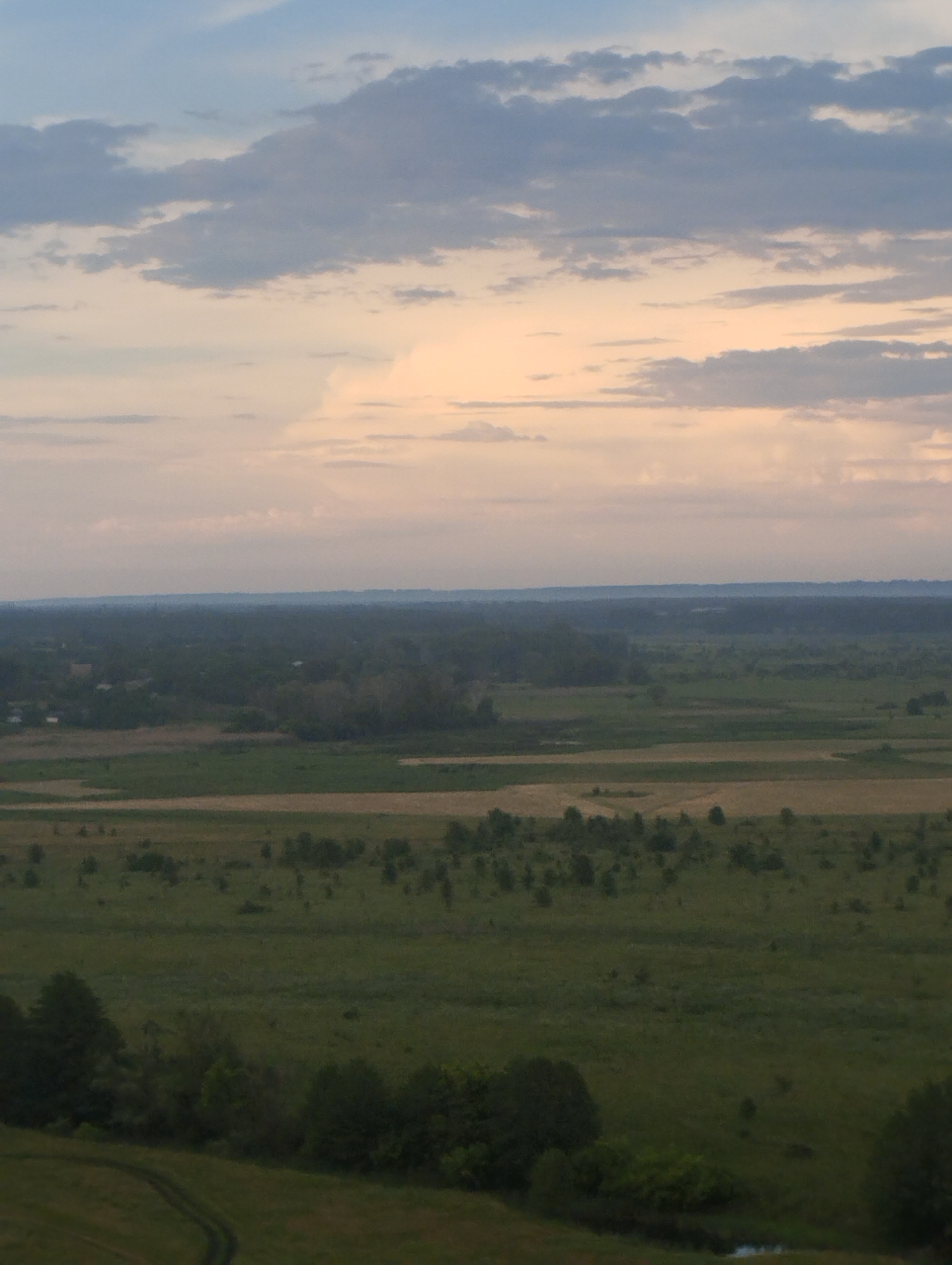 A wide, panoramic view captures a vast rural landscape under a cloudy sky. The sky transitions from a pale blue at the top to soft oranges and pinks near the horizon, suggesting either sunrise or sunset. Below the sky, a distant range of hills is shrouded in a light haze, appearing as a dark, indistinct band. The middle ground is a patchwork of fields, some appearing as green meadows with scattered trees and shrubs, and others with lighter brown tones, possibly indicating cultivated land or dry grass. A village or cluster of houses can be glimpsed amidst the trees in the far distance, marked by small, light-colored structures. The foreground features a dense expanse of green grass with scattered trees and the winding path of a dirt road. The overall impression is one of a serene, natural environment at dawn or dusk.