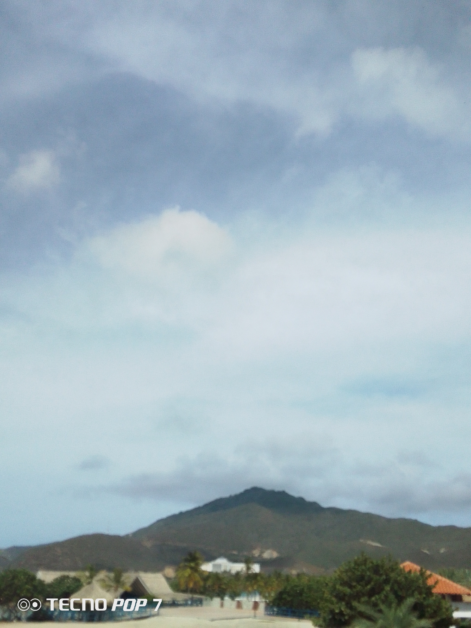 A distant mountain looms over the coastal town of Pedro González, Venezuela. The sky is predominantly cloudy, but interspersed with patches of blue. In the foreground, there are several low structures with thatched roofs, possibly beachside accommodations or recreational facilities. Trees and greenery are visible, adding to the coastal landscape.