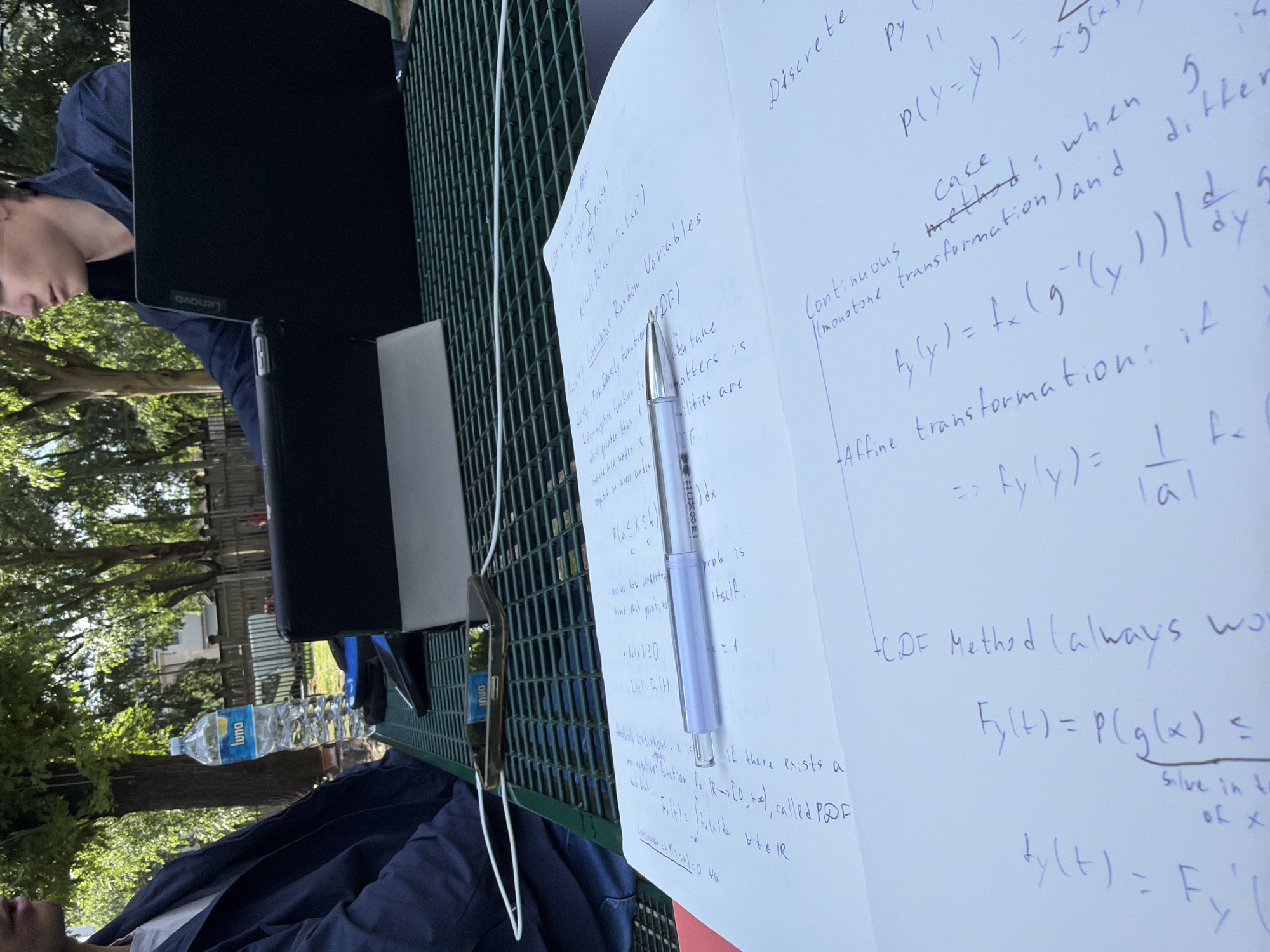 The image appears to be taken outdoors, likely at a park or garden setting, possibly in Milan, Italy, due to the presence of an Italian brand of bottled water. The scene includes a person wearing a blue shirt, possibly studying or working, with a Lenovo laptop, a notebook, a pen, a smartphone, and a bottle of "Luna" water on a green metal table or bench. The notebook contains handwritten notes on probability and statistics, including concepts like discrete and continuous random variables, PDF, and CDF. The focus is on the academic materials and the person involved in studying them in an outdoor environment.