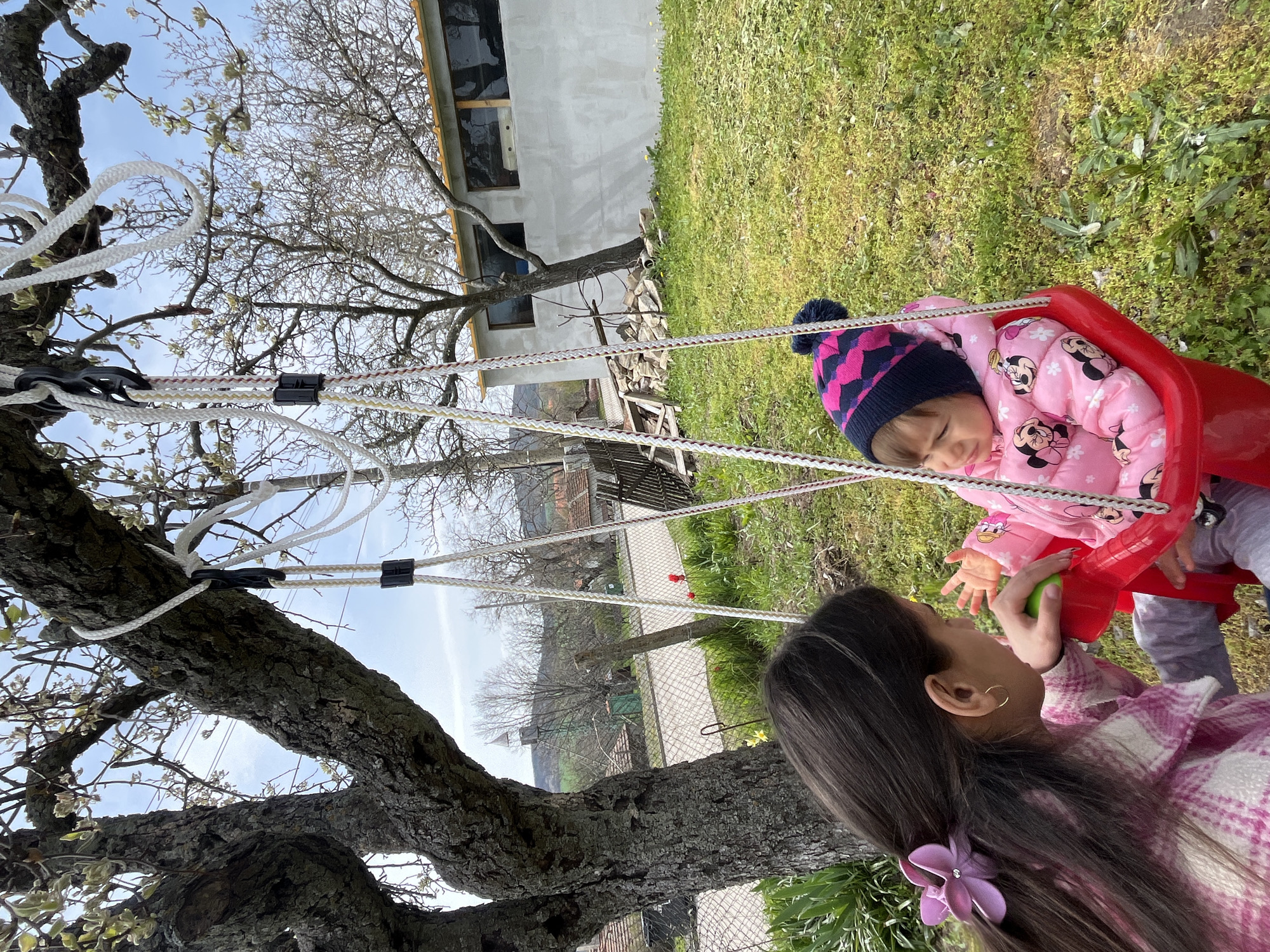 The image shows a young girl in a red swing being pushed by an older girl in a yard. The younger girl is wearing a pink jacket with Mickey Mouse designs and a blue and pink hat. The older girl has long brown hair with a purple flower clip. The swing is hanging from a large tree, and the background shows a grassy area with a house in the distance. The location is likely Kosten, Bulgaria, as indicated.