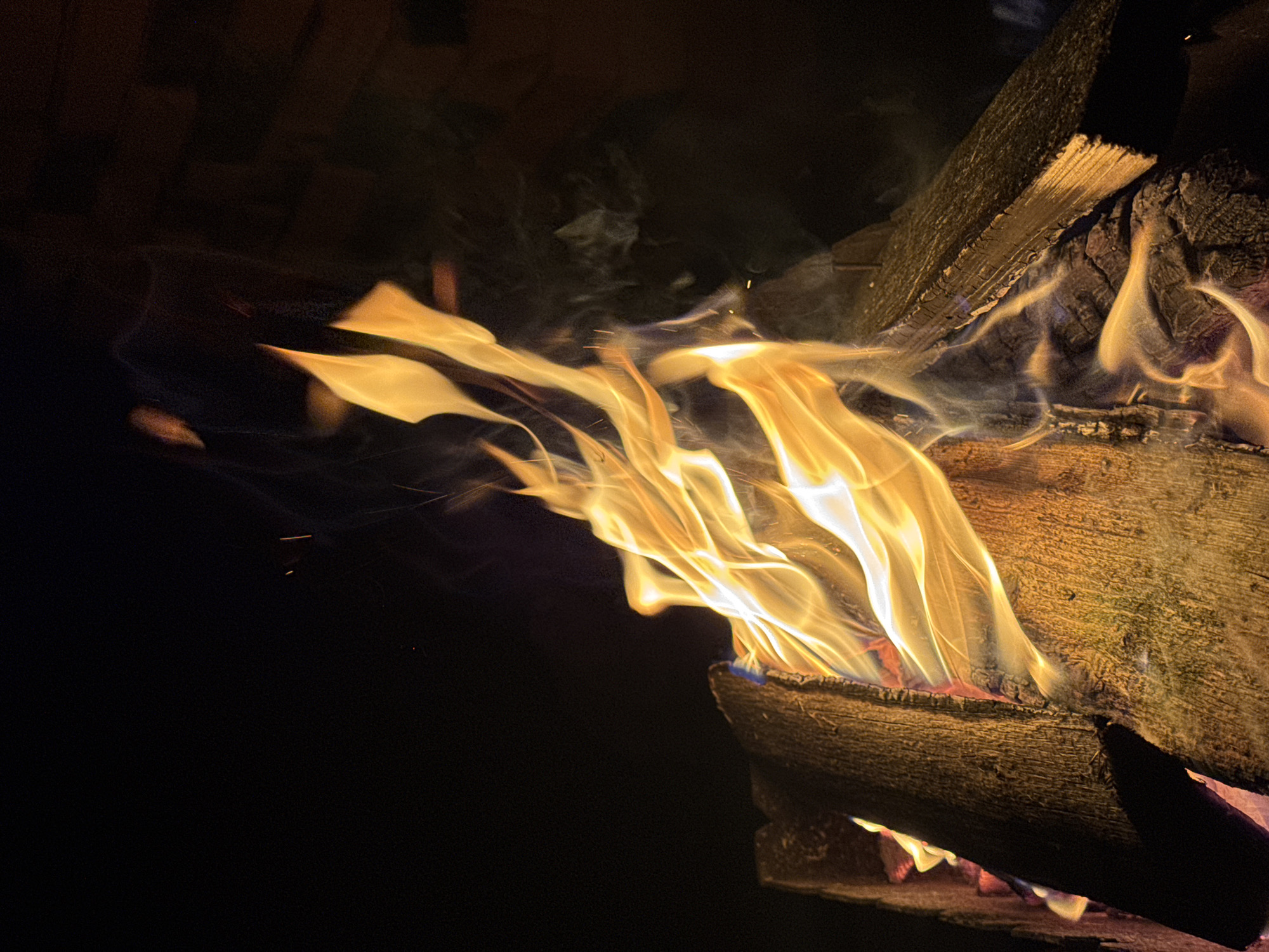 The image shows a close-up of a campfire at night, with flames and burning logs. The background is dark, with a hint of a brick structure on the right side. There are no people visible in the image. The fire is actively burning, with vibrant orange and yellow flames licking upwards. Several pieces of wood are visible, some appearing charred and others still catching fire. Wisps of smoke are also present, contributing to the atmosphere. The lighting is primarily from the fire itself, casting a warm glow on the wood and creating dramatic shadows. The overall impression is one of a cozy and natural setting, likely outdoors. It is not possible to determine the exact location within the city of Maksimovo, Russia, from the image alone, as it does not contain any recognizable landmarks or identifying features of the city.