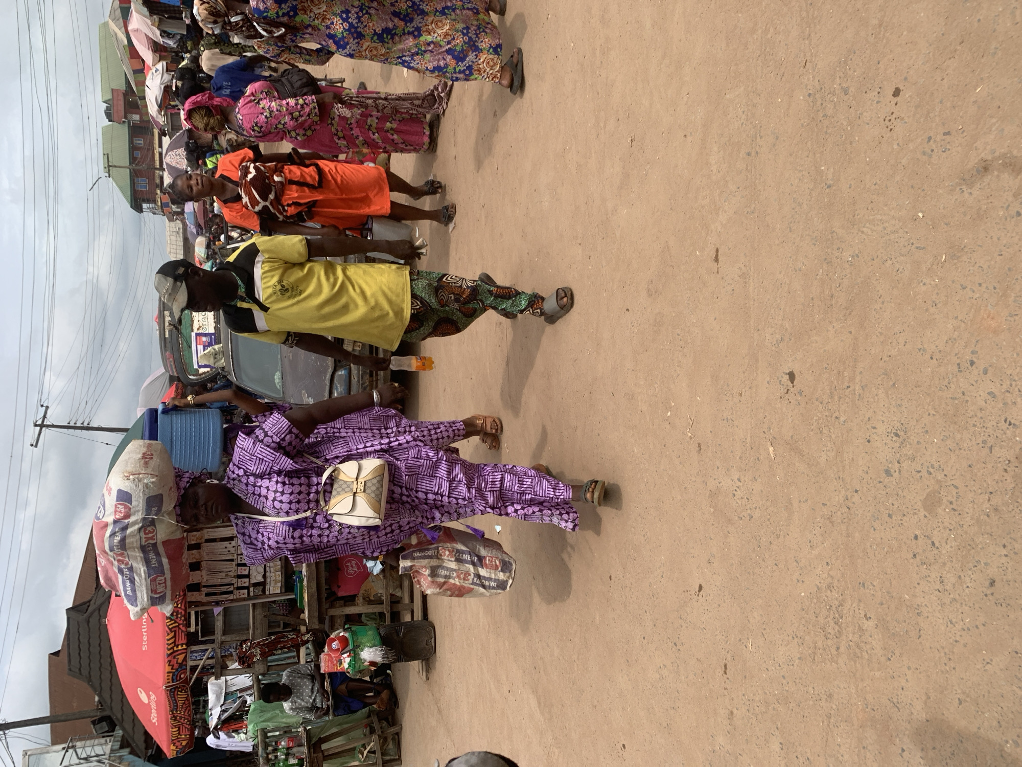 A medium shot captures an outdoor street scene in Shagamu, Nigeria, with a focus on several individuals walking and standing amidst market stalls. In the foreground, a woman with a light-skinned complexion walks to the left, carrying a large sack of cement on her head and a plastic bag in her right hand. She is adorned in a vibrant purple traditional outfit with a matching headscarf and a cream and gold purse slung over her shoulder. To her right, a man wearing a yellow polo shirt with black trim and patterned green and black trousers walks to the right. He holds an orange beverage bottle in his right hand. Behind them, other people are visible in the midground. A woman in an orange dress is walking to the right, and another woman in a pink and black floral dress is also walking to the right. The background reveals a bustling market environment with numerous stalls, umbrellas, and a partially visible vehicle. The ground is dusty and uneven. The sky is overcast with visible power lines stretching across the frame.