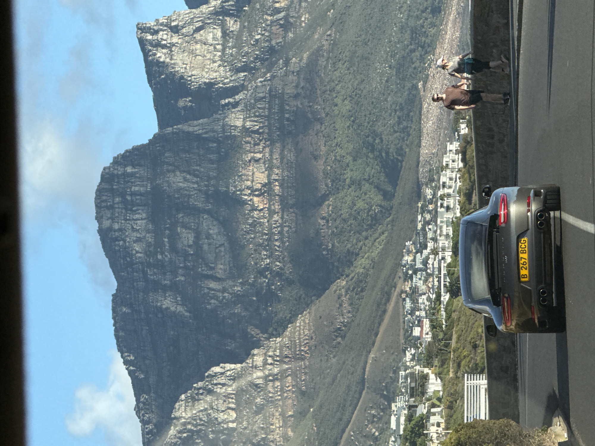 The image shows a view of Table Mountain in Cape Town, South Africa. In the foreground, a gray car with the license plate "B 267 BCG" is parked on the roadside. Two people are standing near the roadside, with a townscape visible in the valley below. Table Mountain dominates the background under a clear blue sky.