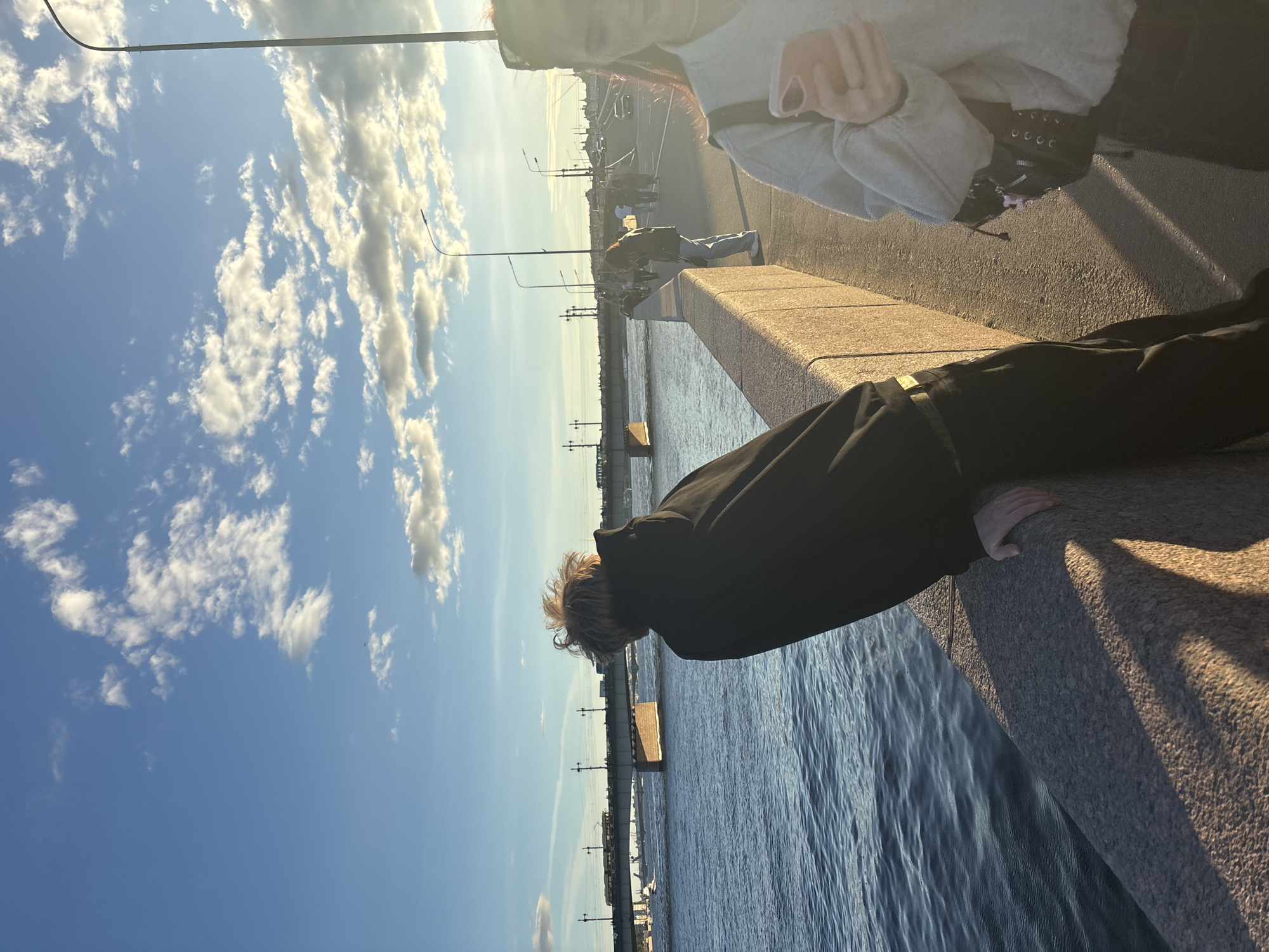 The image depicts a waterfront scene in Saint Petersburg, Russia, under a partly cloudy blue sky during what appears to be late afternoon or early evening, given the warm, low-angle sunlight casting long shadows. In the foreground, a person with light-colored, tousled hair, wearing a black hooded sweatshirt and dark pants, leans against a stone embankment, looking out over a wide body of water. The water is a deep blue, with gentle ripples.
Across the water, a long bridge stretches from left to right, supported by stone piers. Various forms of transportation, including what appear to be trams or light rail trains, are visible on the bridge, as well as traffic and pedestrians. The bridge itself has streetlights and is a prominent feature of the cityscape.
To the right of the person leaning on the embankment, another individual is visible from the chest down, holding a pink smartphone and wearing a light gray hooded sweatshirt and dark pants. This person appears to be walking away from the camera. The embankment is constructed of large, rough-hewn granite blocks. The overall atmosphere is one of urban leisure, with people enjoying the waterfront and the city's architectural grandeur.