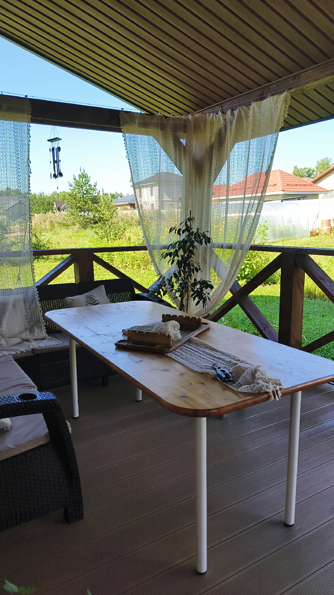 A wooden table with white legs sits on a brown deck, with a wicker couch to the left and a potted plant on the table. The scene appears to be an outdoor covered patio in a rural area, possibly a dacha in Sokury, Russia. The background shows a green lawn, trees, and houses, suggesting a residential setting. A wind chime hangs from the ceiling.