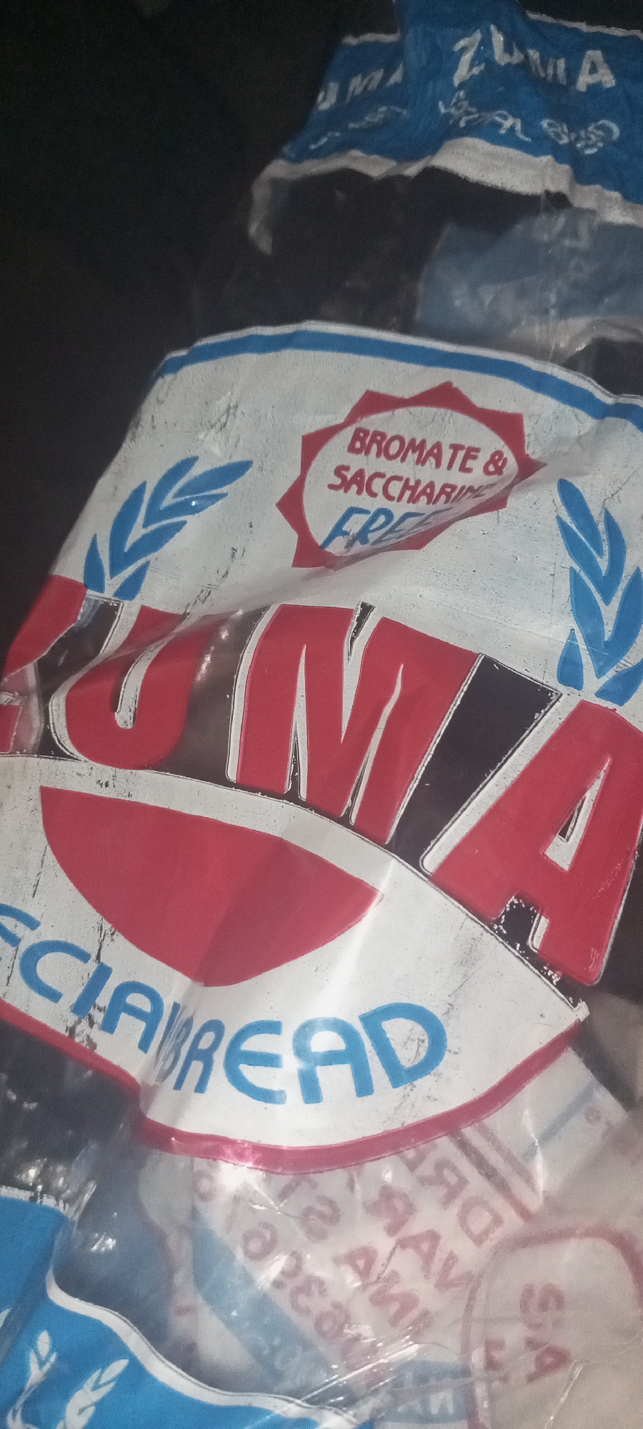 The image shows a crumpled bread bag with the brand name "ZUMA" printed in large red letters with a black outline. Above the brand name, a red starburst shape contains the text "BROMATE & SACCHARIN FREE" in red and blue. Blue wheat stalks are visible on either side of the brand name. The words "SPECIAL BREAD" are visible in blue text along the bottom of the bag. The bag appears to be made of plastic and is translucent in some areas, revealing other items underneath. The background is dark and out of focus, suggesting the image was taken in low light conditions or with shallow depth of field. The location is listed as Unknown, Unknown.