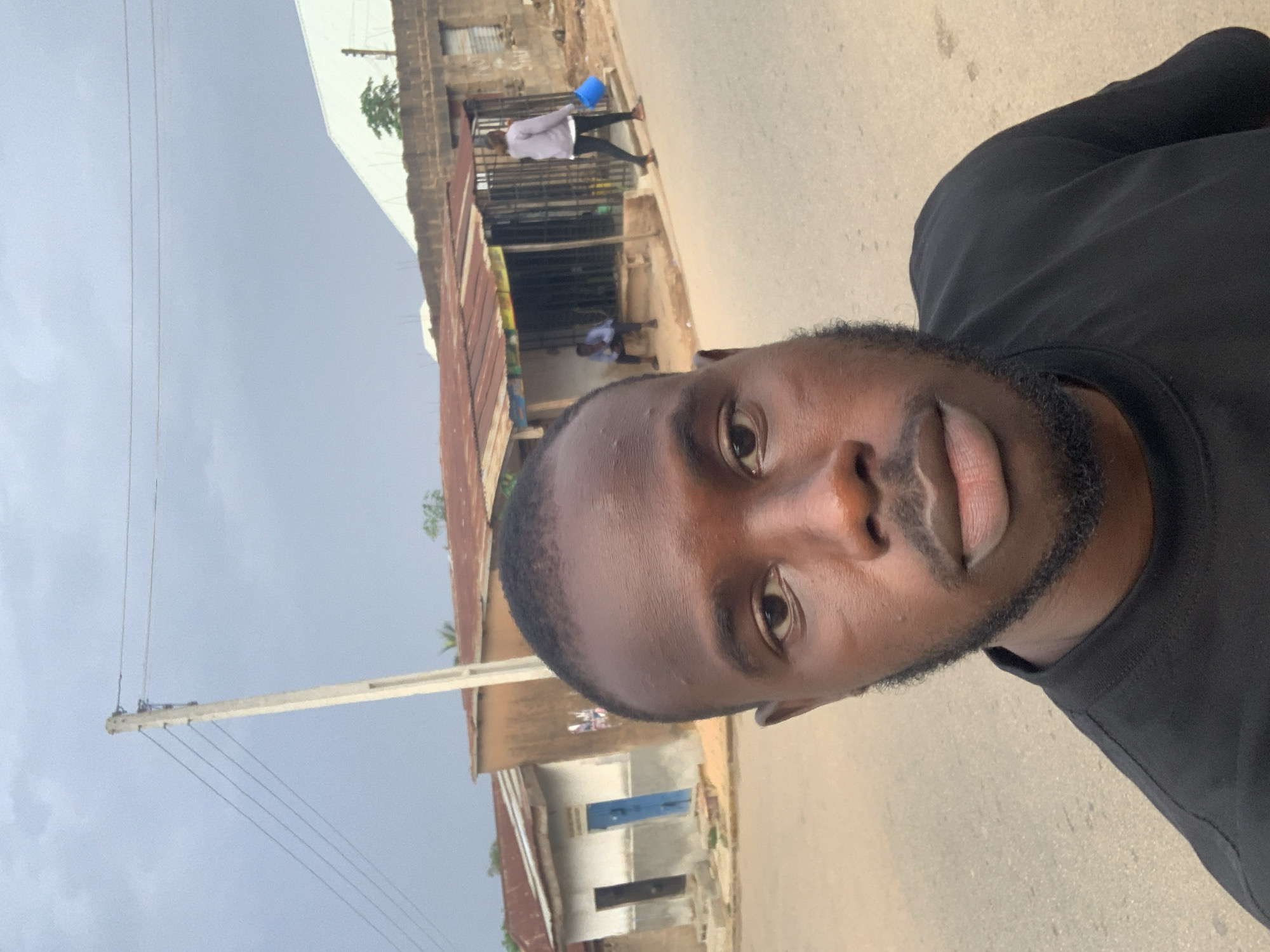 A close-up, slightly angled selfie of a dark-skinned man with a short, neatly trimmed beard, wearing a black shirt. The background shows a street scene in Jos, Nigeria, with buildings of varying architectural styles and conditions. One building has a rusty corrugated iron roof. People can be seen in the background, including one individual carrying a blue bucket. A utility pole with wires is visible on the left side of the frame. The sky is overcast.